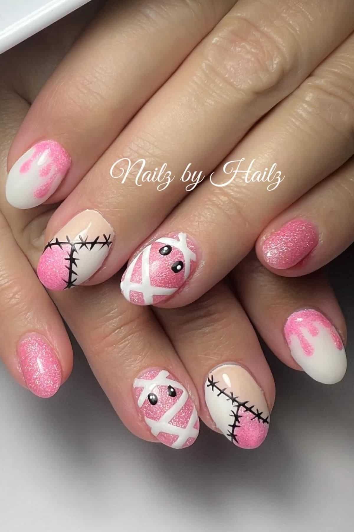 Hands with pink and white nail art featuring glitter, dripping patterns, stitched designs, and mummy faces with black eyes.