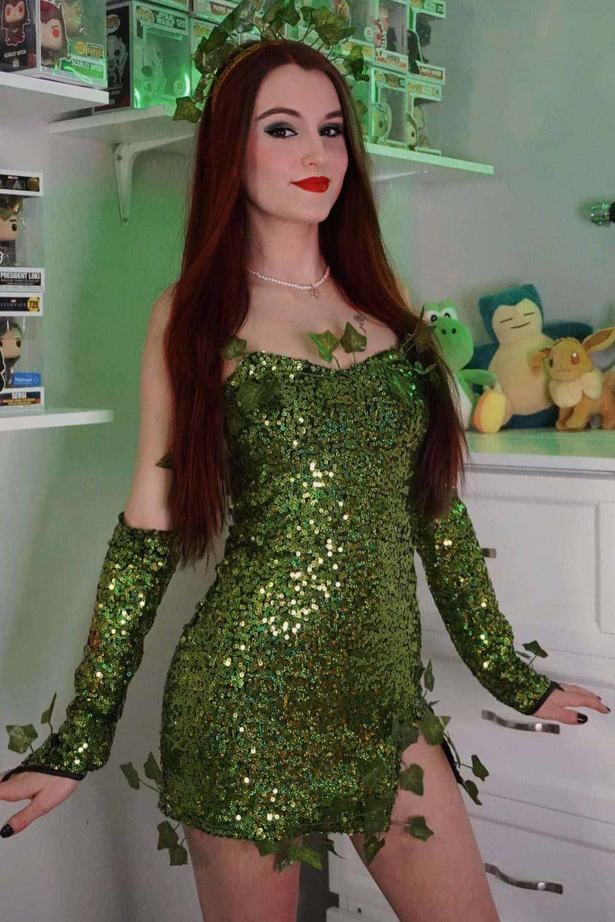 Person dressed in a green, sequin-covered costume with leaf accents, long red hair, and red lipstick, standing indoors near shelves with toys and collectibles.