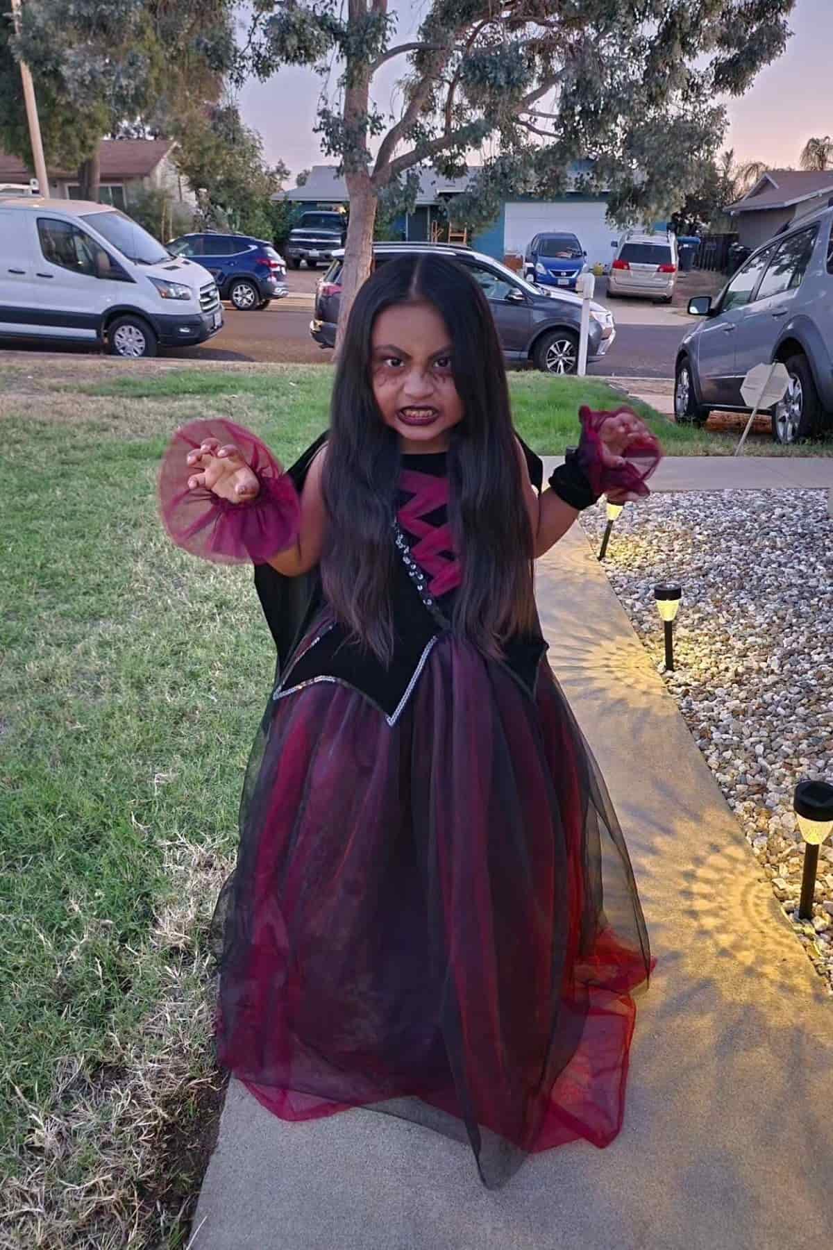 Child in a black and red vampire costume stands on a sidewalk with hands raised, baring fangs. Cars and houses are visible in the background.