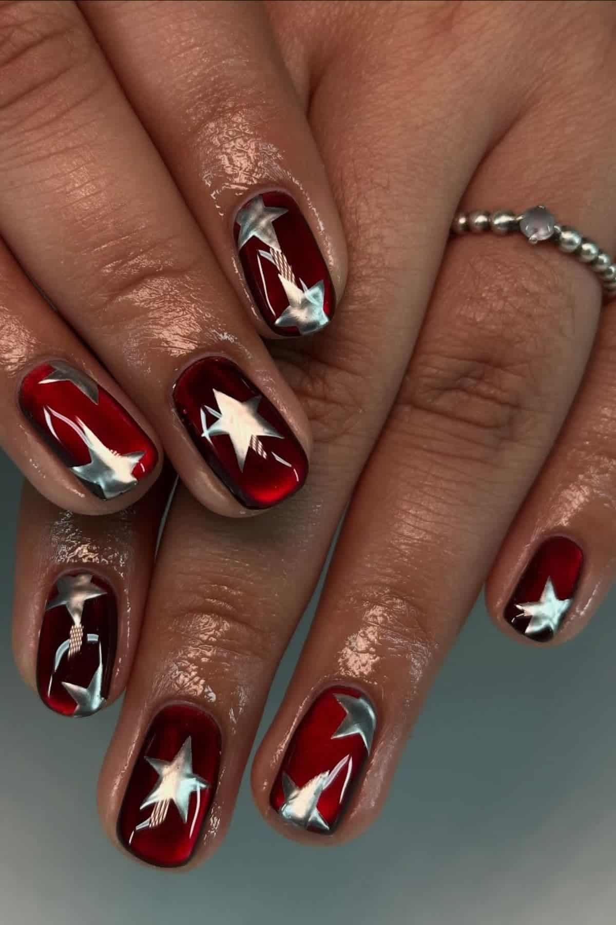A hand with glossy red nails featuring metallic silver star designs on each nail. The person wears a beaded silver ring on the pinky finger.