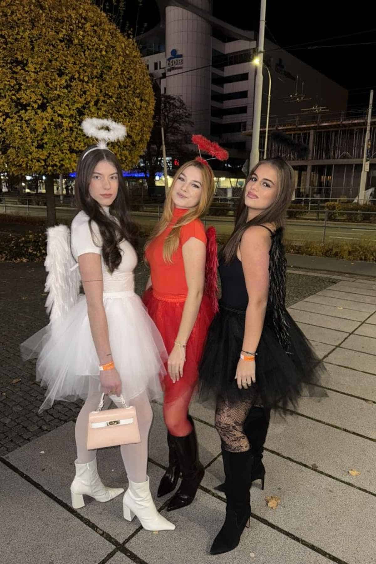 Three young women in angel and devil costumes pose outdoors at night; one in white, one in red, one in black, each with matching wings and halos, standing on a sidewalk near a tree and buildings.