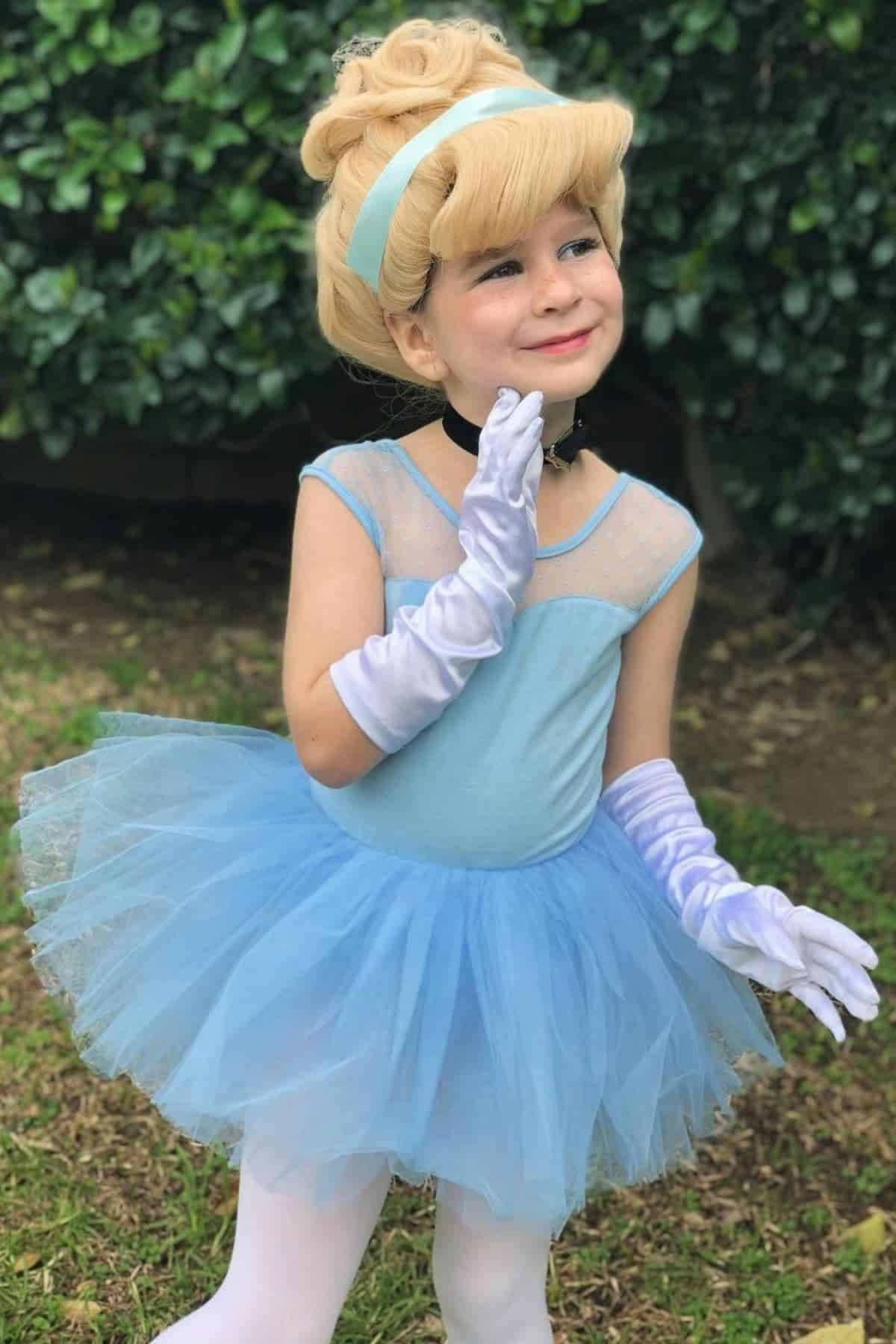 A young child in a blue tutu dress, white gloves, and a blonde wig poses outdoors, dressed as a princess.
