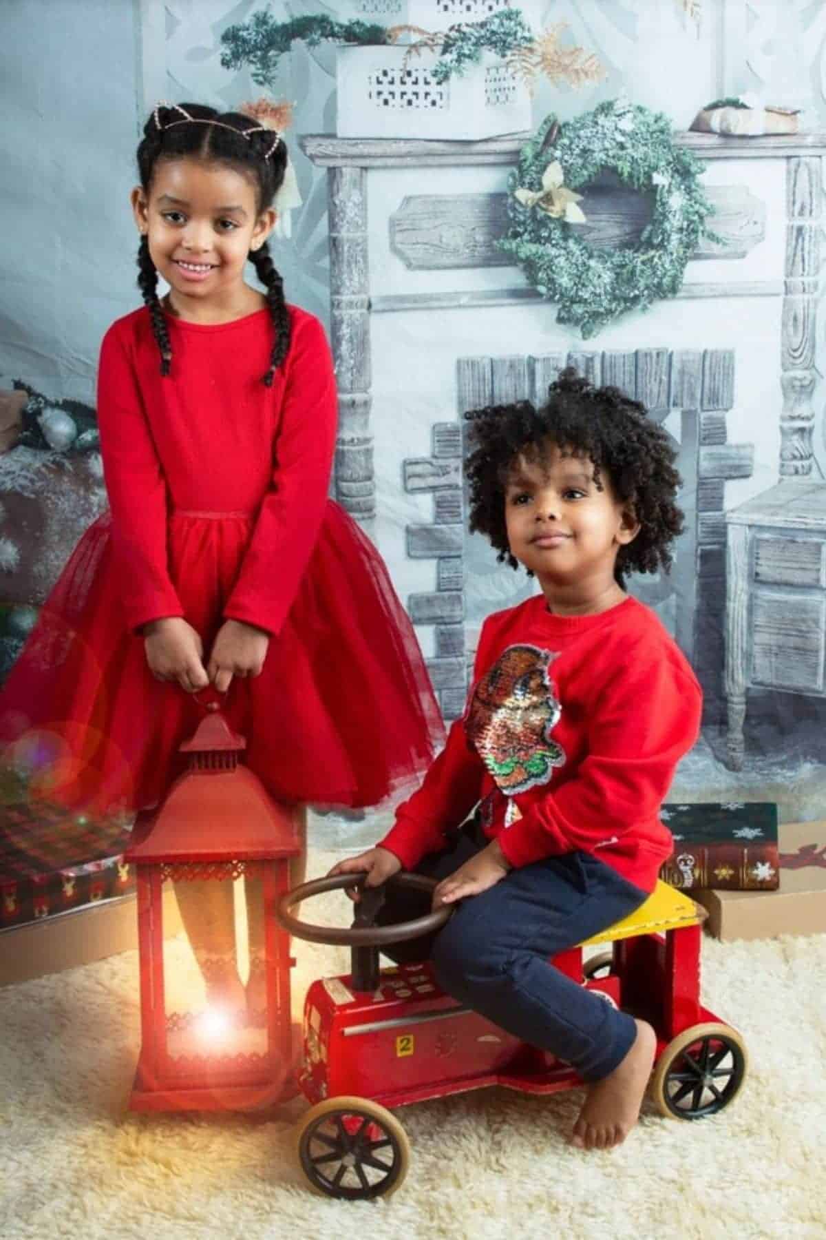Two children in red outfits pose indoors; one stands holding a red lantern, the other sits on a red toy car. A festive, winter-themed backdrop decorates the background.