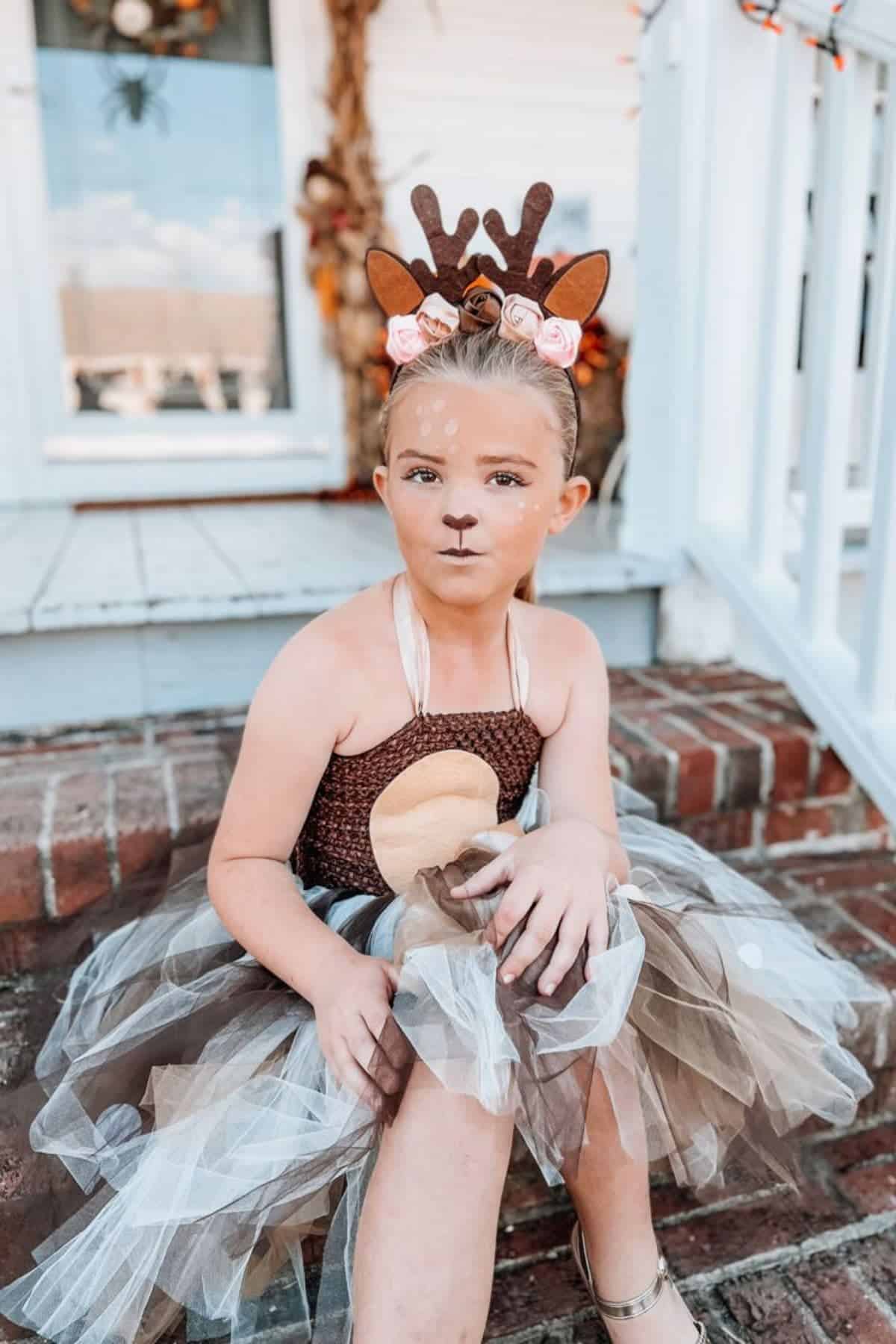 Young child in a brown and tan tutu dress sits on brick steps, wearing a headband with deer antlers and flowers, with face painted to resemble a deer.