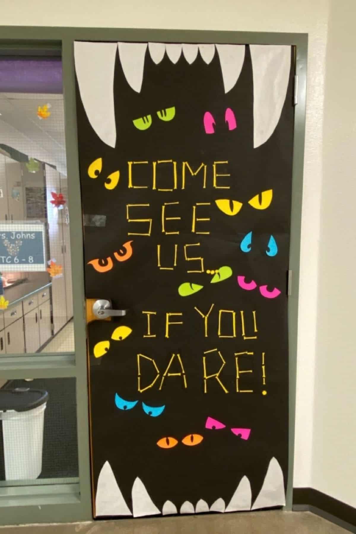 Classroom door decorated with large monster teeth, colorful eyes, and the words “COME SEE US IF YOU DARE!” written in yellow letters.