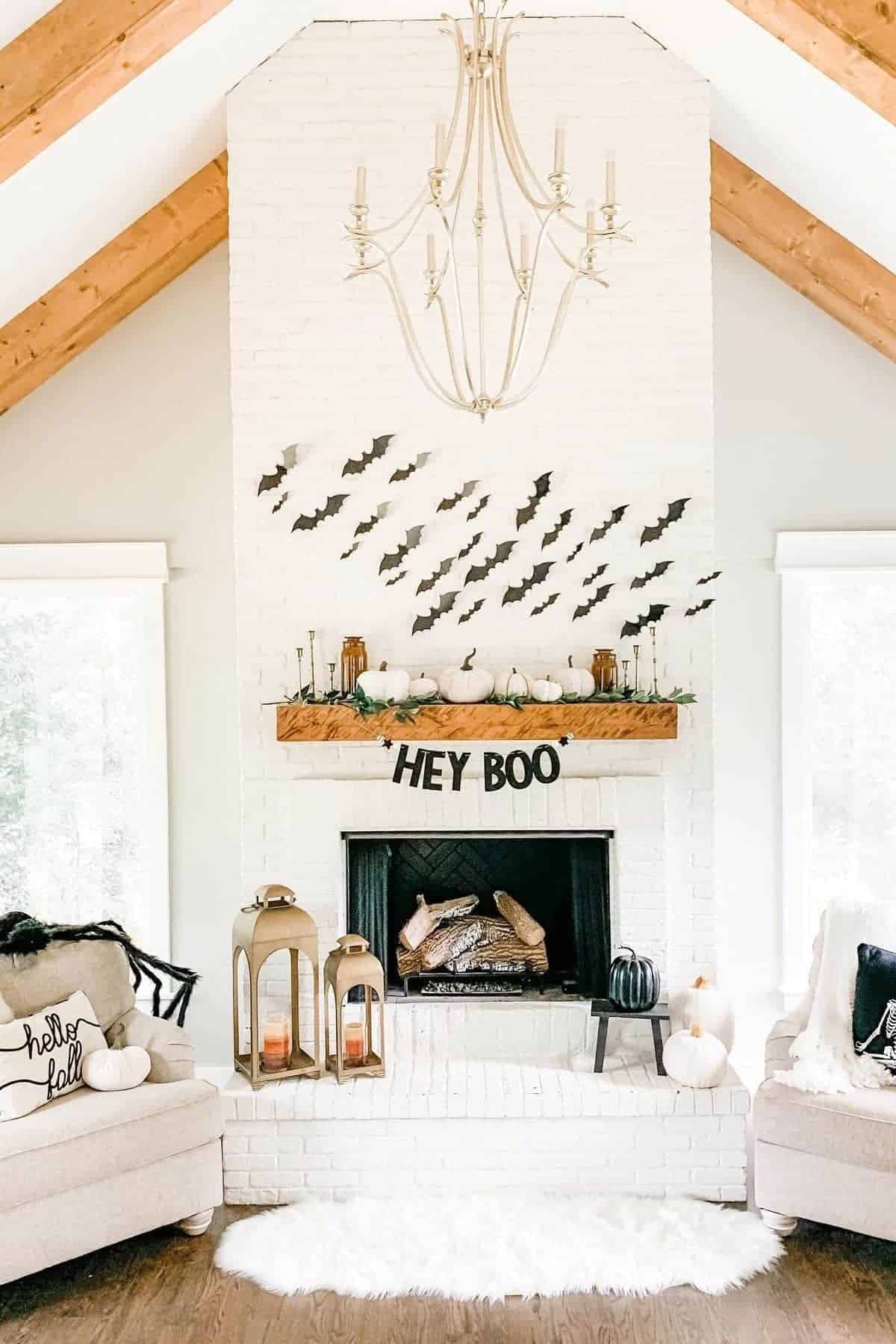 A white brick fireplace decorated with black paper bats, small pumpkins, a “HEY BOO” banner, and lanterns. Two beige chairs with fall-themed pillows flank the fireplace.