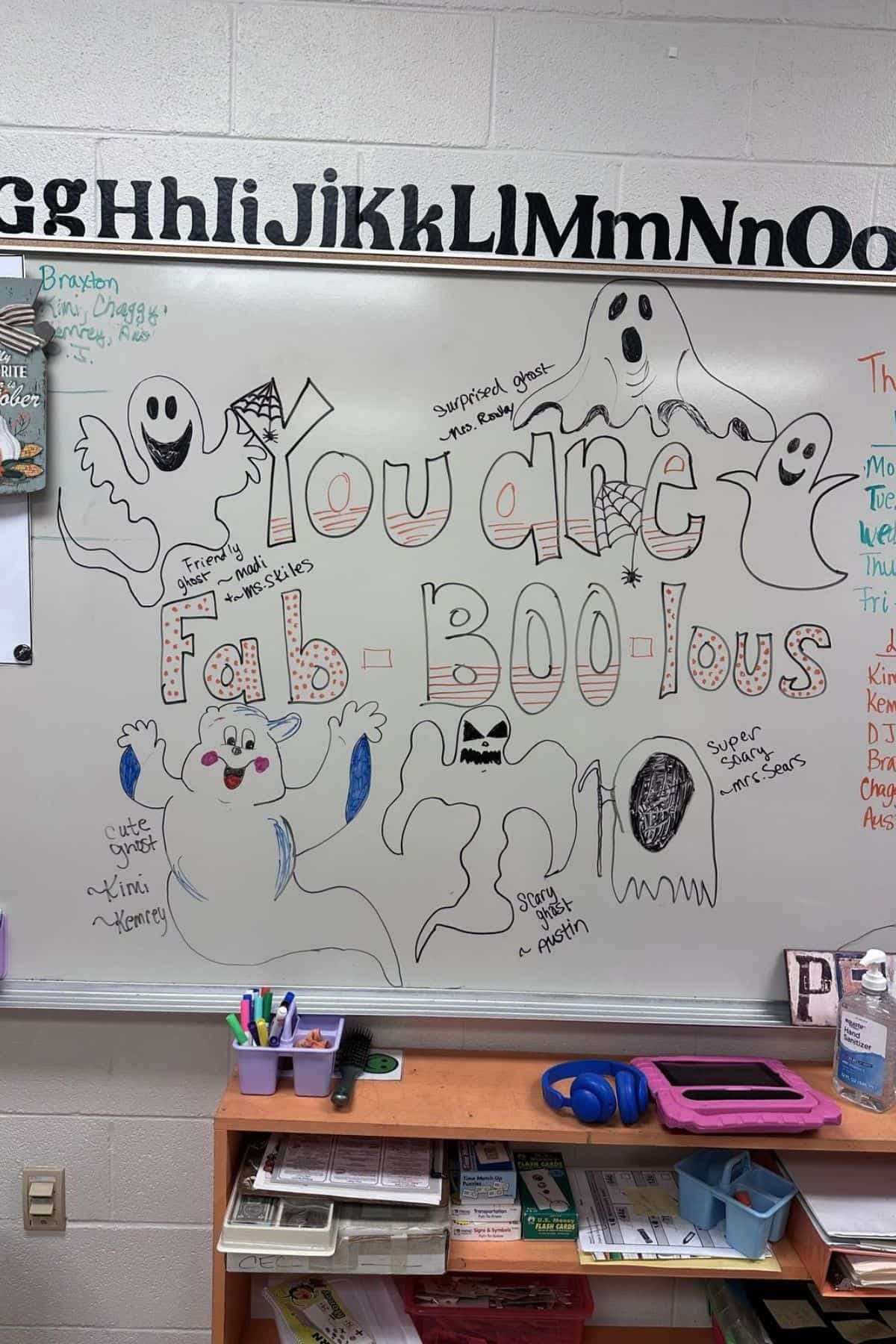 Whiteboard with “You are fab-BOO-lous” in colorful bubble letters, surrounded by drawings of cartoon ghosts and messages from students. Classroom supplies sit on a wooden shelf below.