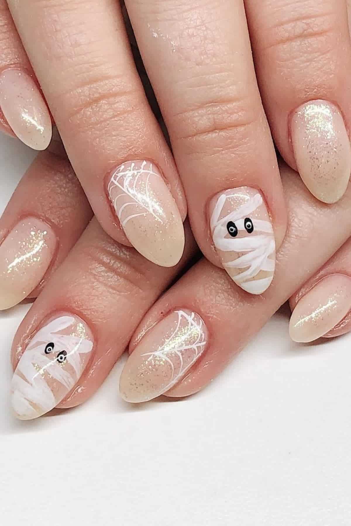 Close-up of hands with almond-shaped nails featuring glittery nude polish, white spider web designs, and two accent nails painted with a mummy pattern and black eyes.