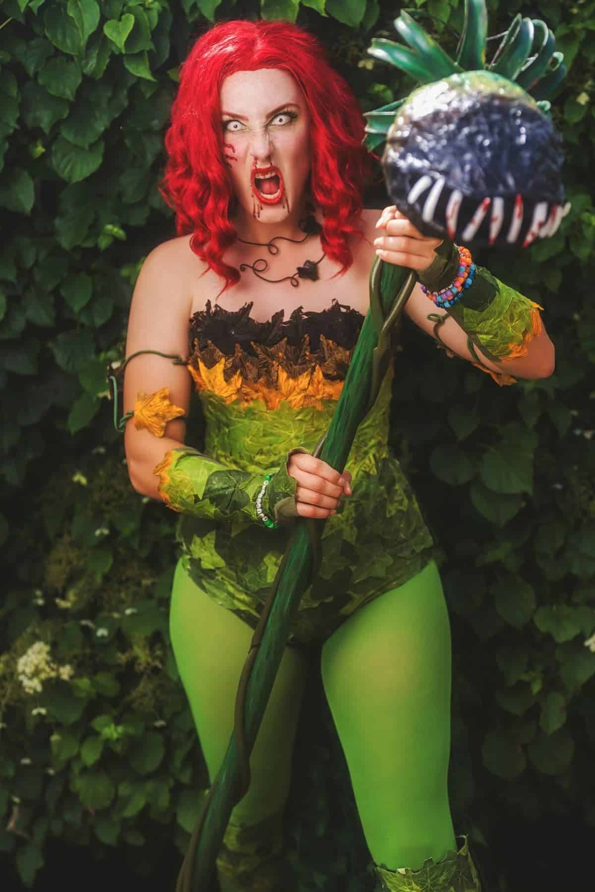 Person in a green and yellow nature-inspired costume with red hair, holding a staff with a large carnivorous plant prop, standing in front of leafy foliage.