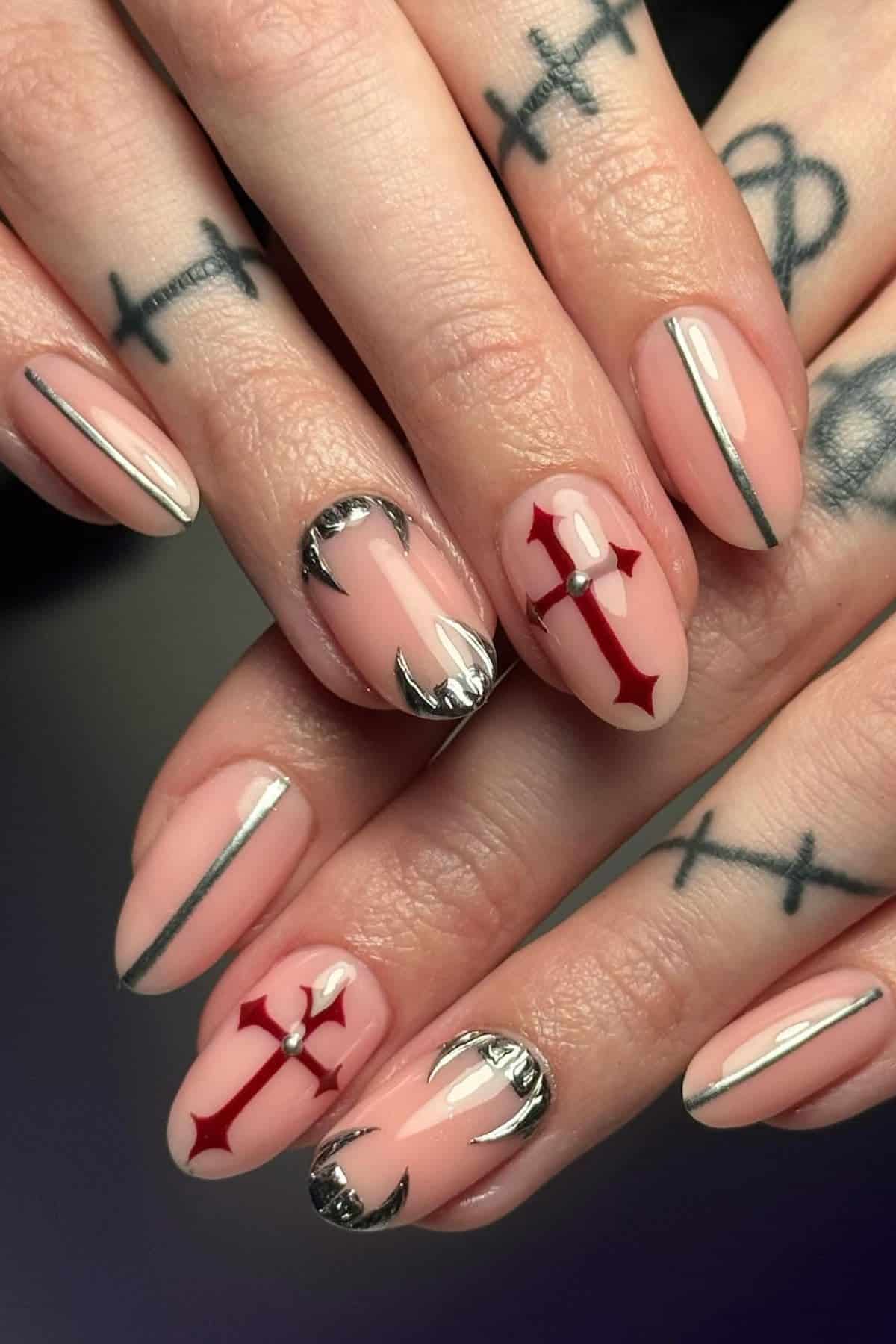 Hands with nude almond-shaped nails featuring red cross designs, silver accents, and thin metallic lines; fingers have bold black cross tattoos.