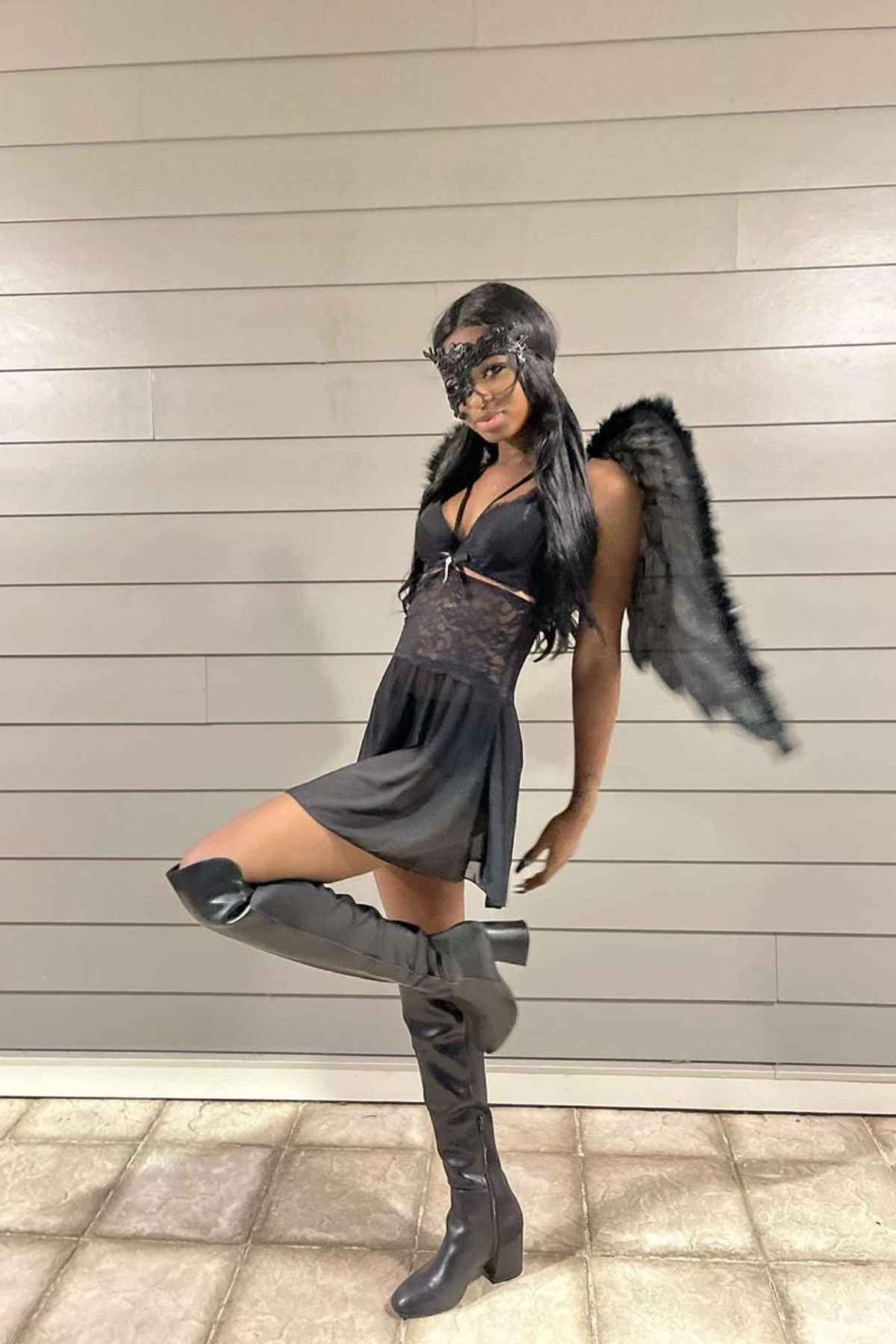 Person wearing a black dress, black boots, black feathered wings, and a black mask poses with one leg raised against a tiled floor and paneled wall.