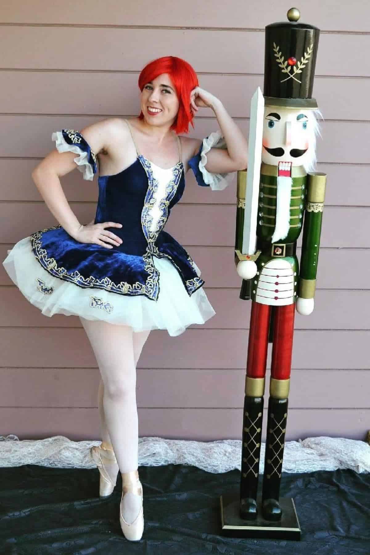 Person in a blue ballerina costume and red wig poses on pointe next to a tall nutcracker statue in front of a wooden wall.