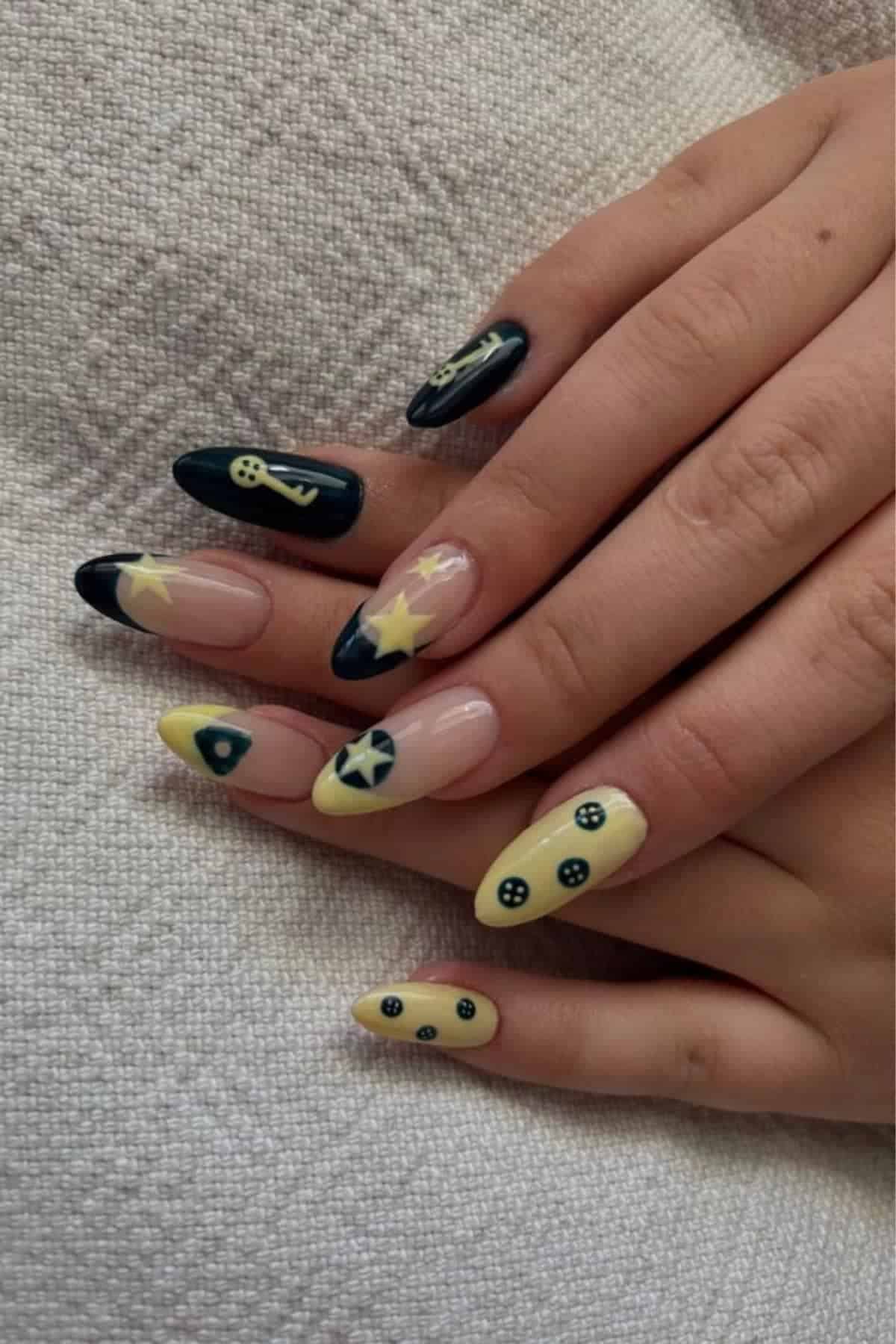 Hands with long almond-shaped nails, painted with yellow and black nautical-themed designs, including anchors, stars, and steering wheels, resting on a textured fabric background.