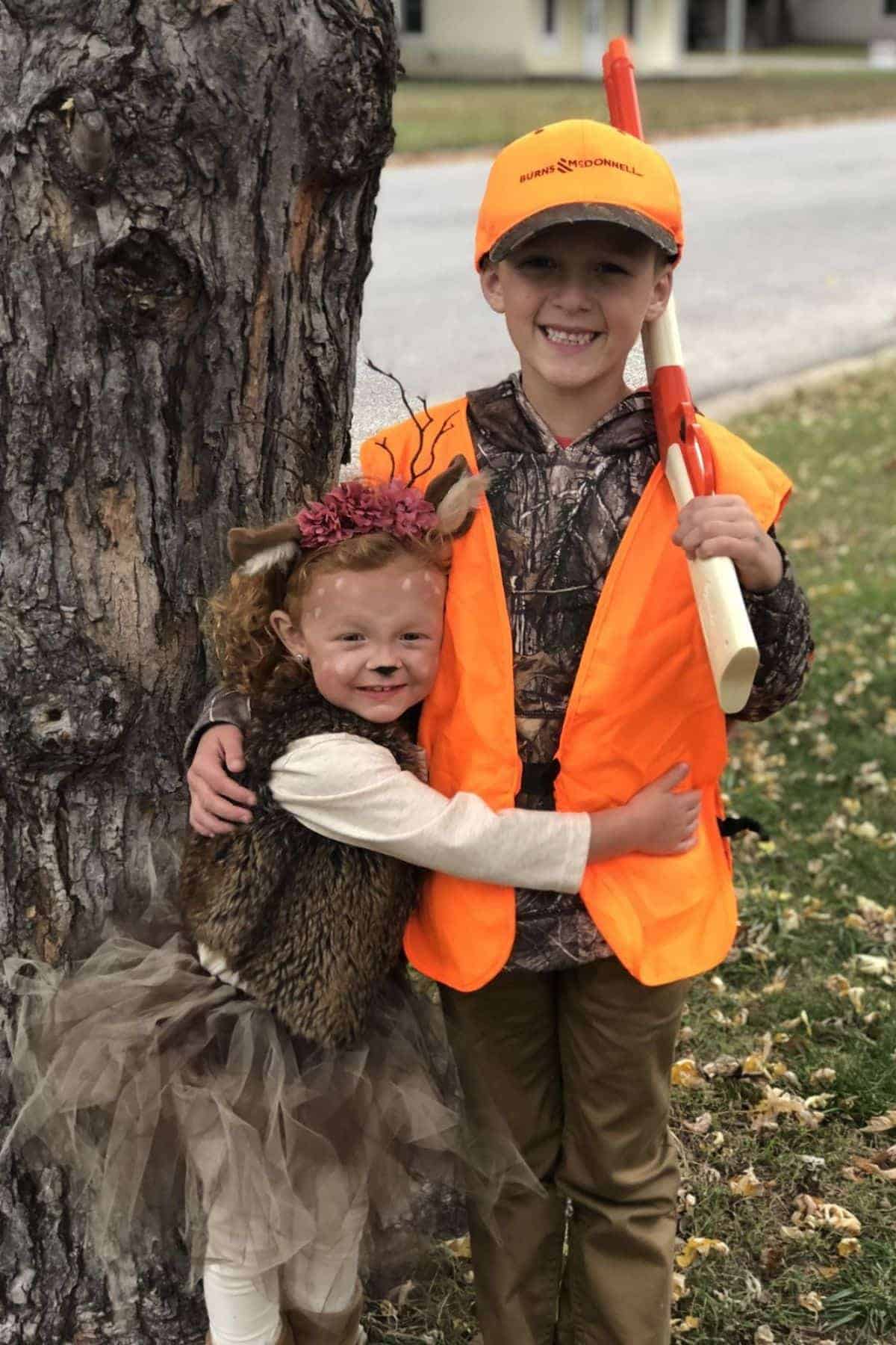Two children in costumes stand by a tree; one is dressed as a deer, the other as a hunter in orange gear with a toy rifle.