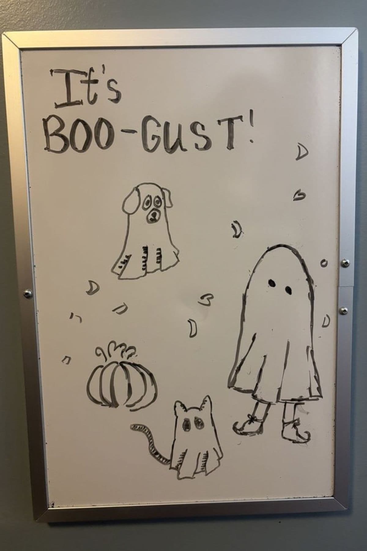 Whiteboard with the phrase “It’s BOO-GUST!” and drawings of a ghost, a sad dog, a pumpkin, and a small cat, surrounded by a few scattered leaves.