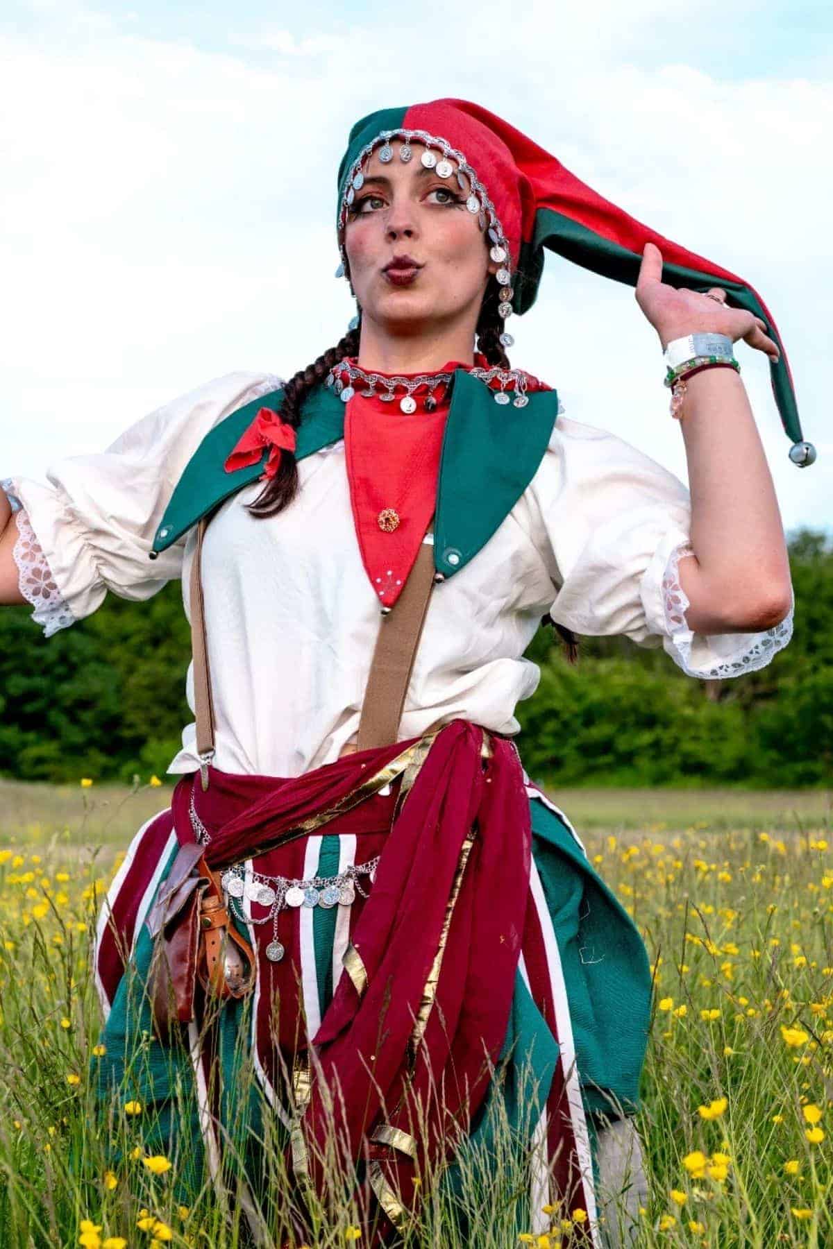 Person wearing a colorful jester costume with a red and green hat stands in a grassy field with yellow flowers.