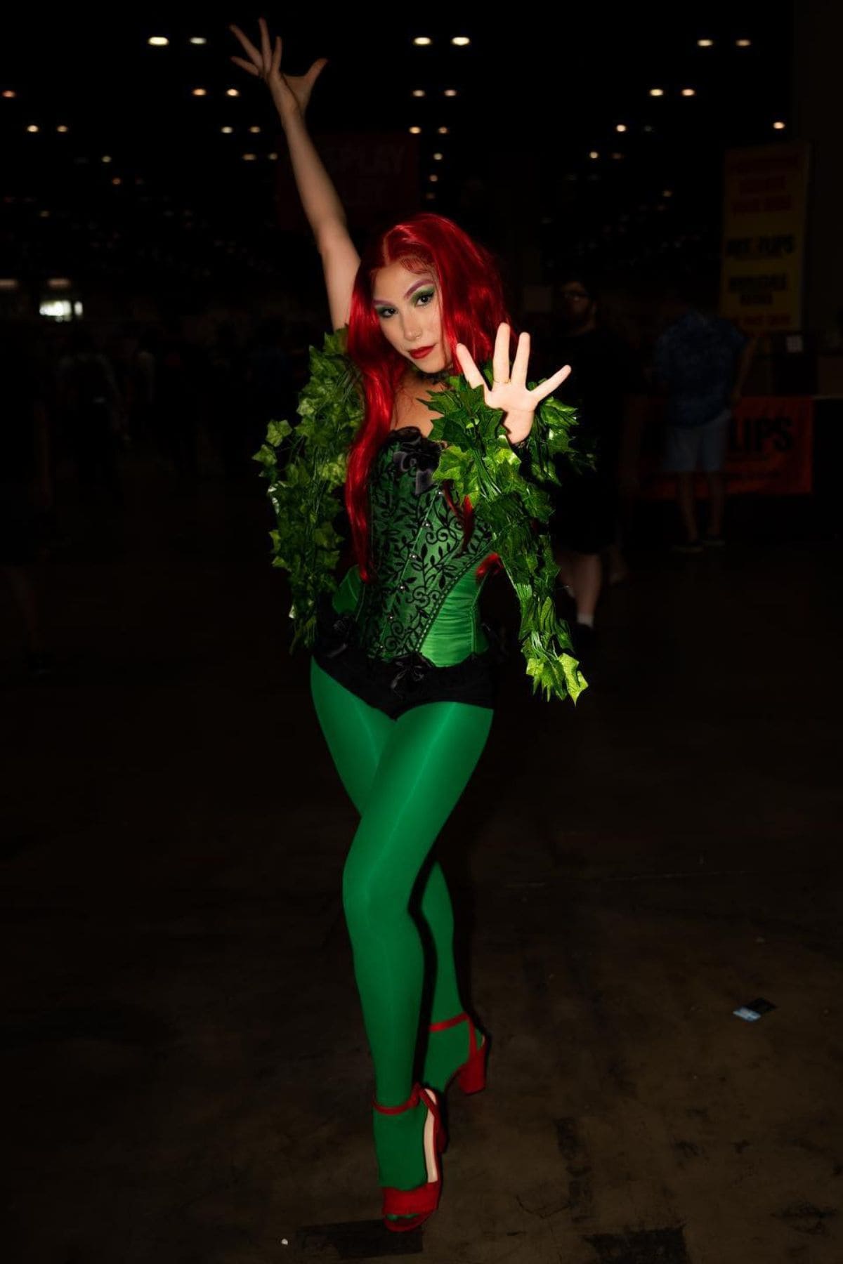 Person in a green corset, leafy accessories, red wig, and green tights poses at a convention, cosplaying as a comic book character.