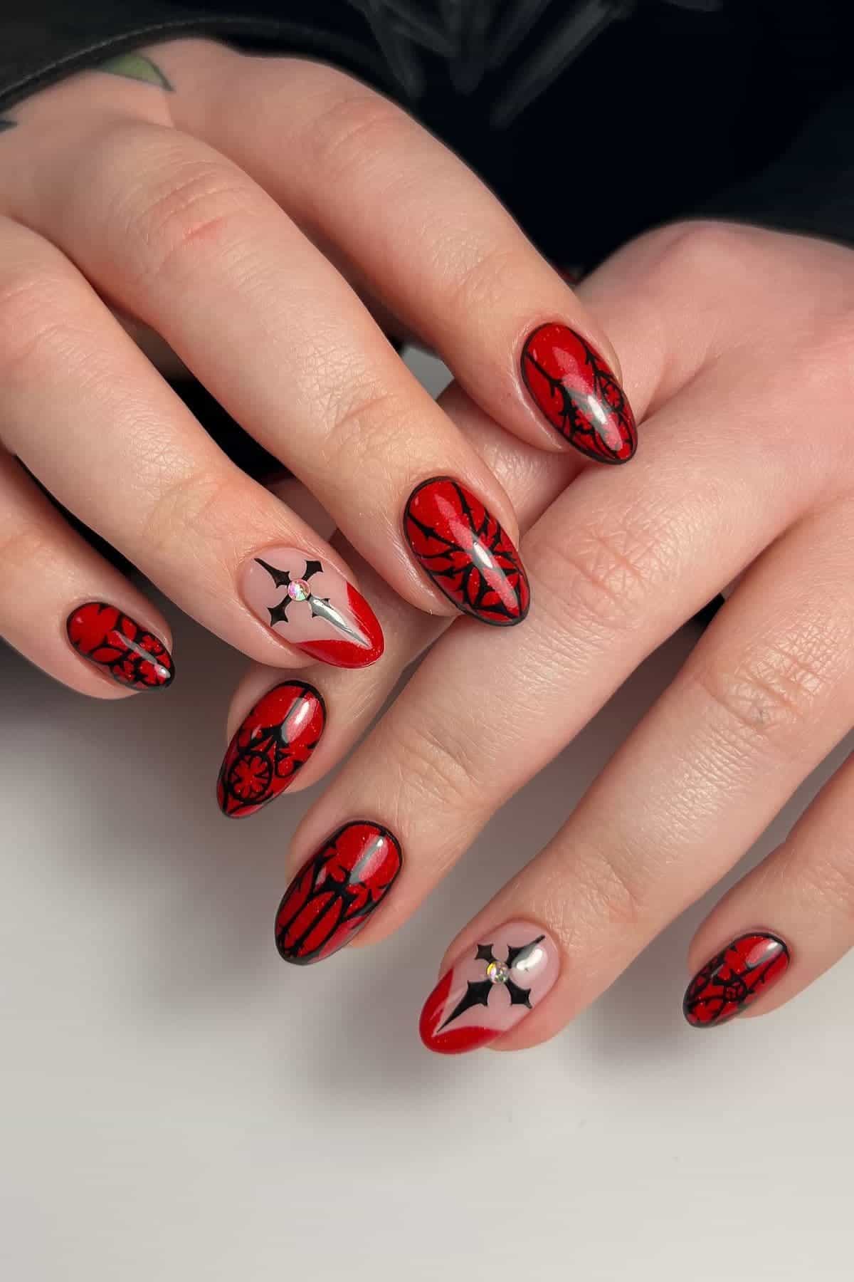 Hands with almond-shaped nails featuring red and black spiderweb nail art, with two nails on each hand decorated with white and black star designs on a nude base.