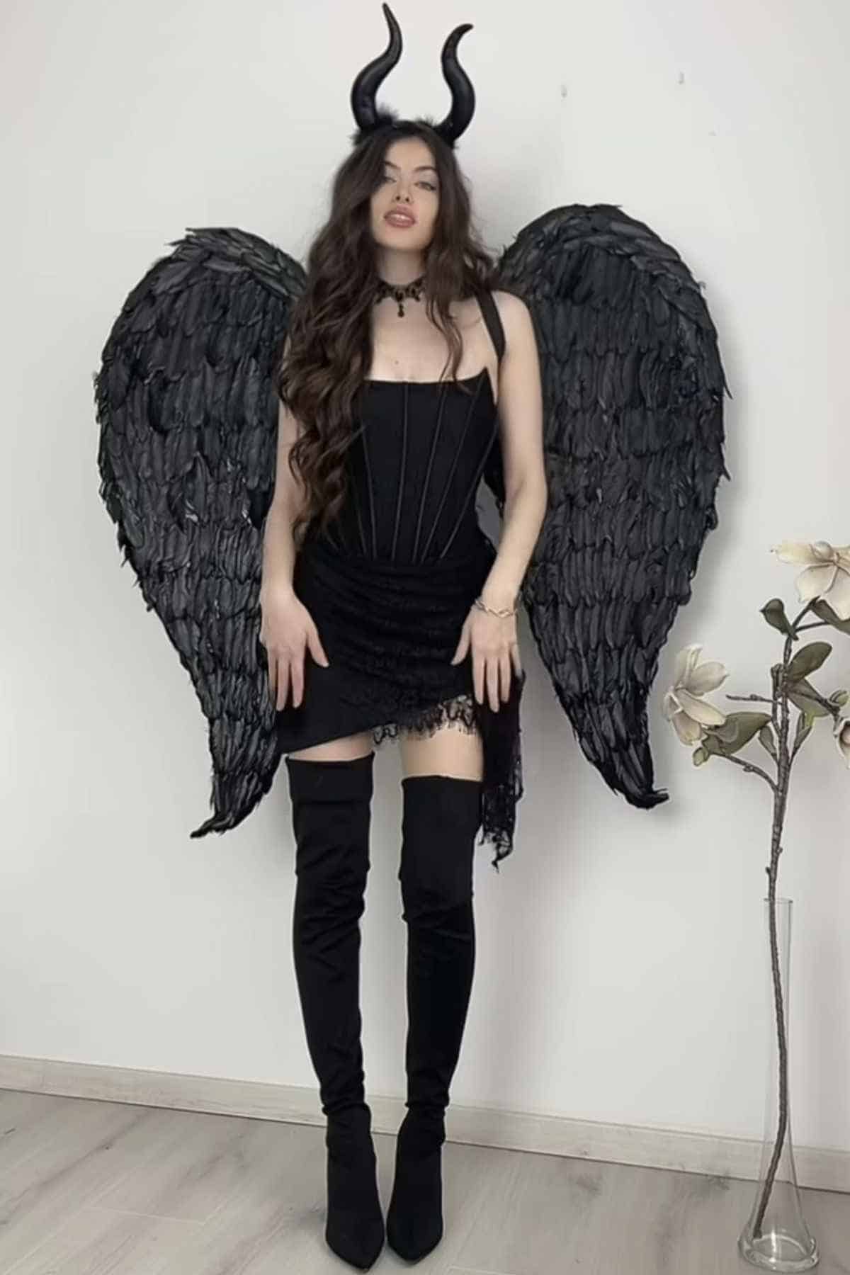A woman in a black dress, black thigh-high boots, black feathered wings, and black horn headband poses indoors beside a white wall and a flower arrangement.