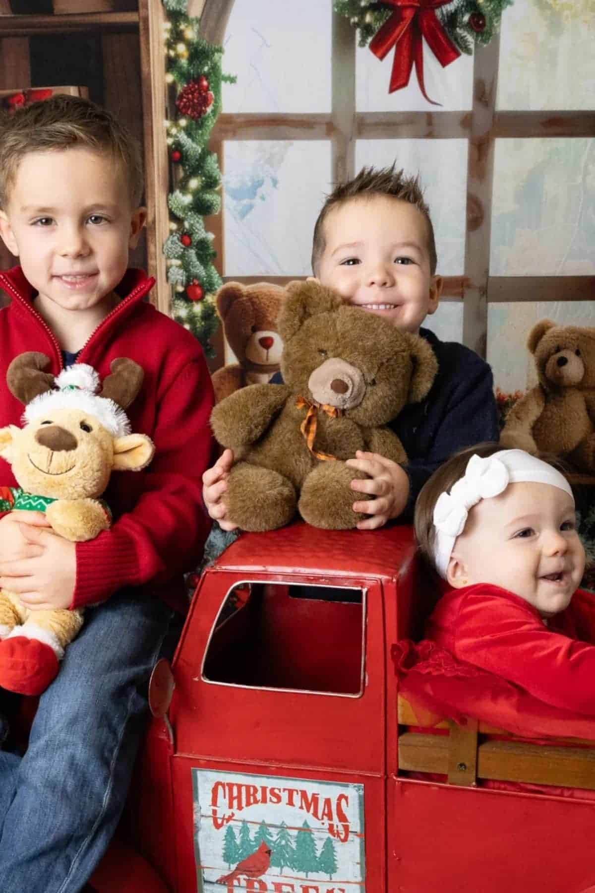 Three young children pose indoors with stuffed animals, sitting on and around a red toy truck decorated for Christmas, with teddy bears and festive holiday decor in the background.