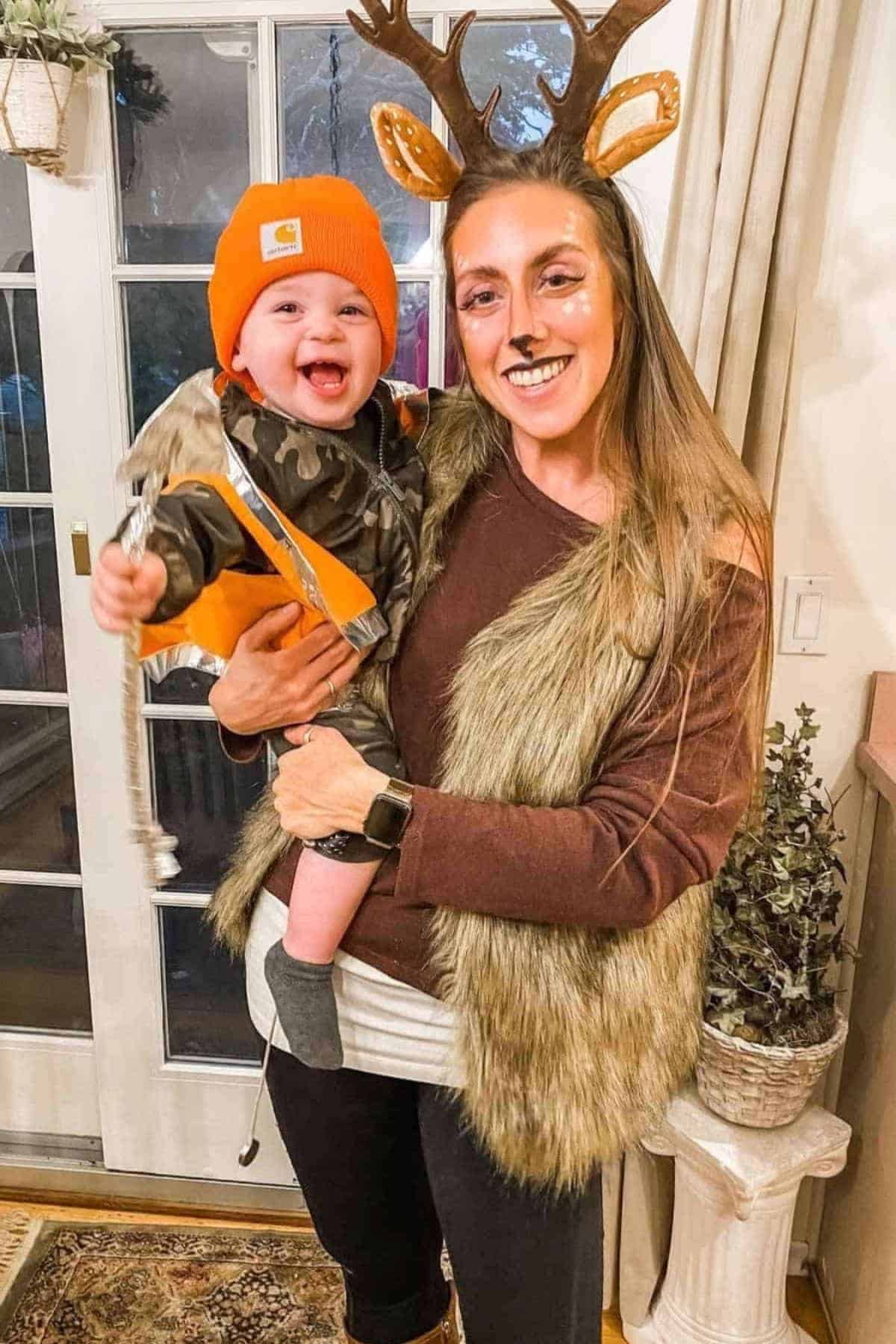 Woman dressed as a deer holds a smiling baby in a camouflage outfit and orange hat inside a home near a glass door.