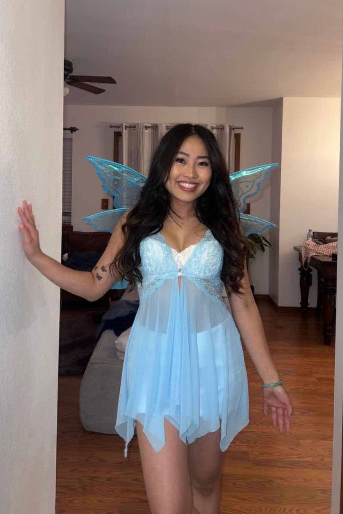 Woman in a light blue fairy costume with wings stands indoors, smiling at the camera with one hand on the wall.
