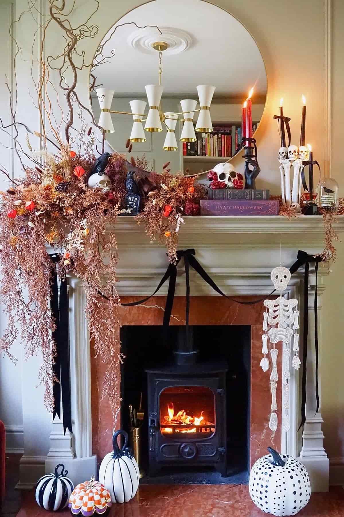 A decorated fireplace with autumn branches, pumpkins, candles, skulls, books, and a skeleton figure; a mirror and chandelier are above, and a fire burns in the wood stove below.