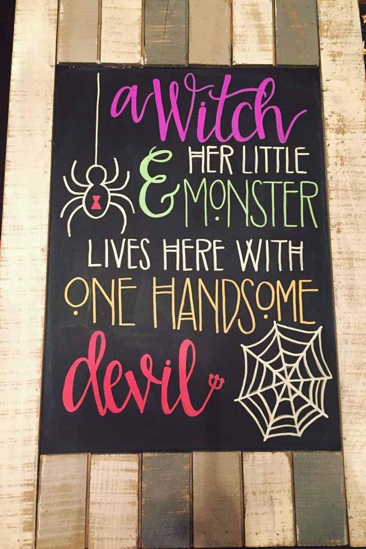 Chalkboard sign with playful Halloween text: “A witch, her little monster, lives here with one handsome devil,” with drawings of a spider and web.