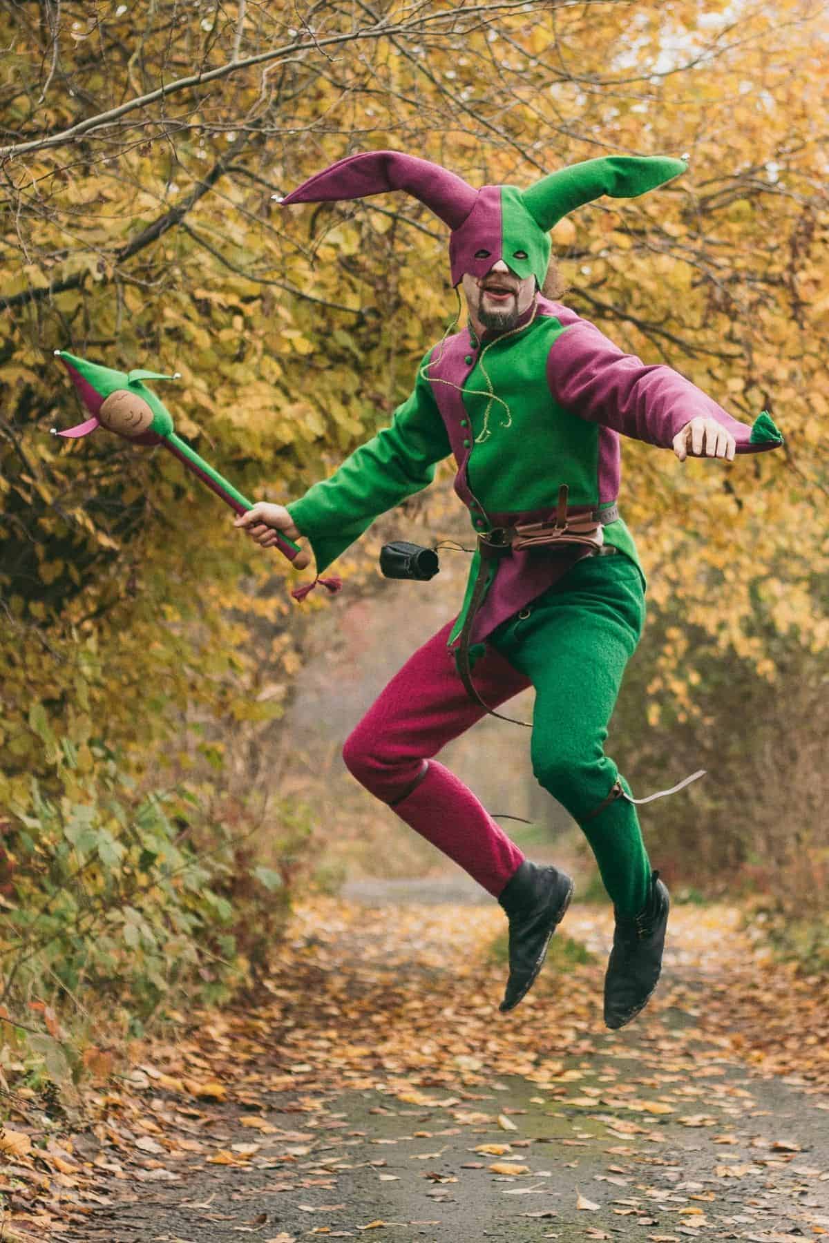 Person in a green and pink jester costume jumps mid-air on a leaf-covered path surrounded by autumn trees, holding a staff.