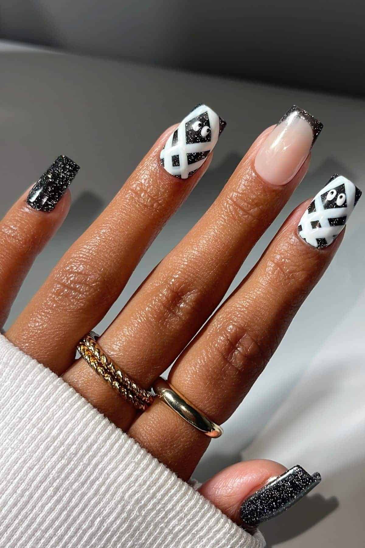 A hand with long square nails featuring black glitter, geometric white lines, and one nude accent nail. The person wears gold rings and a white sweater.