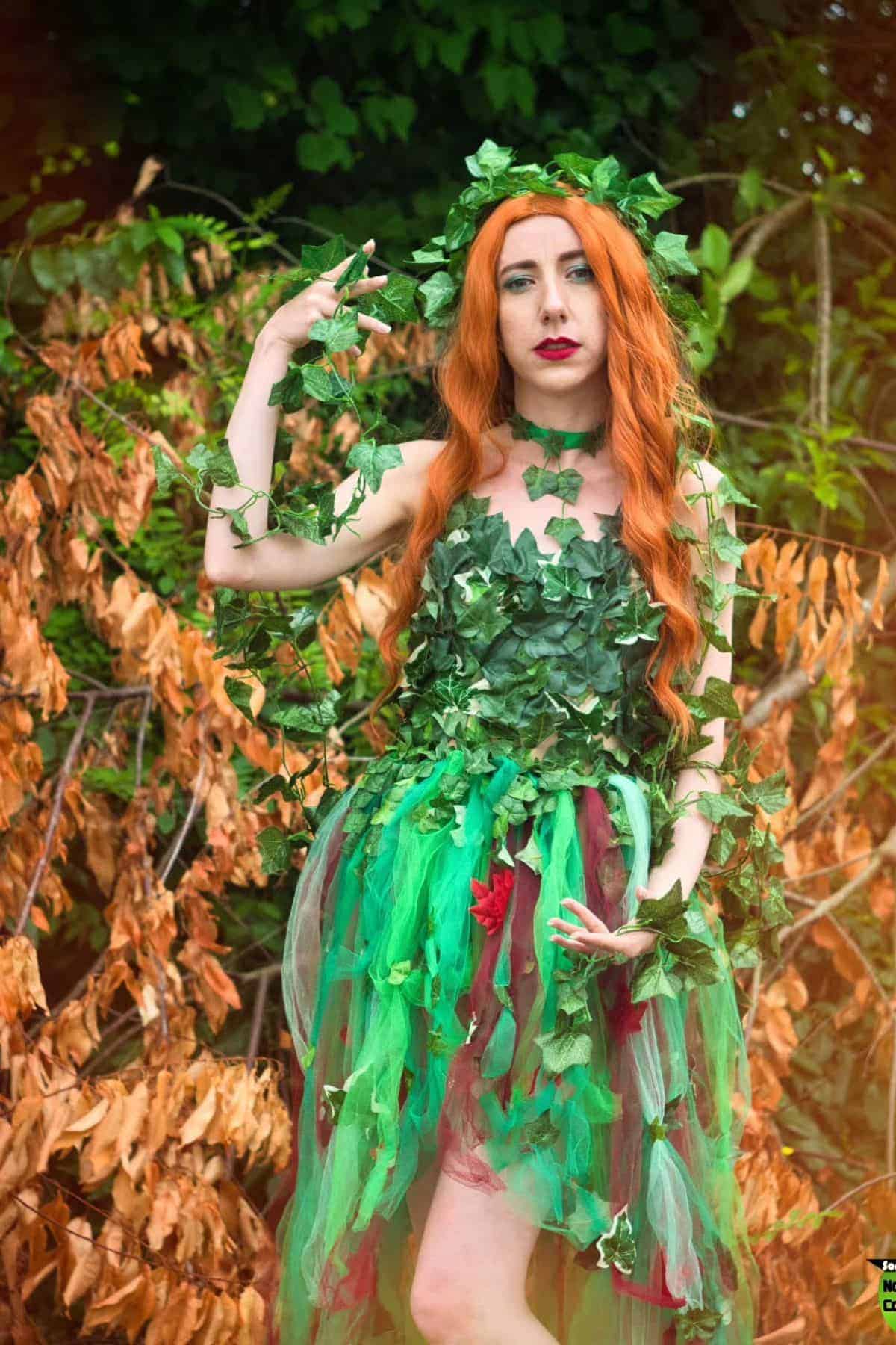 Woman in a leafy green and red costume with long red hair stands outdoors, surrounded by green and brown foliage, wearing a crown of leaves and holding vines.