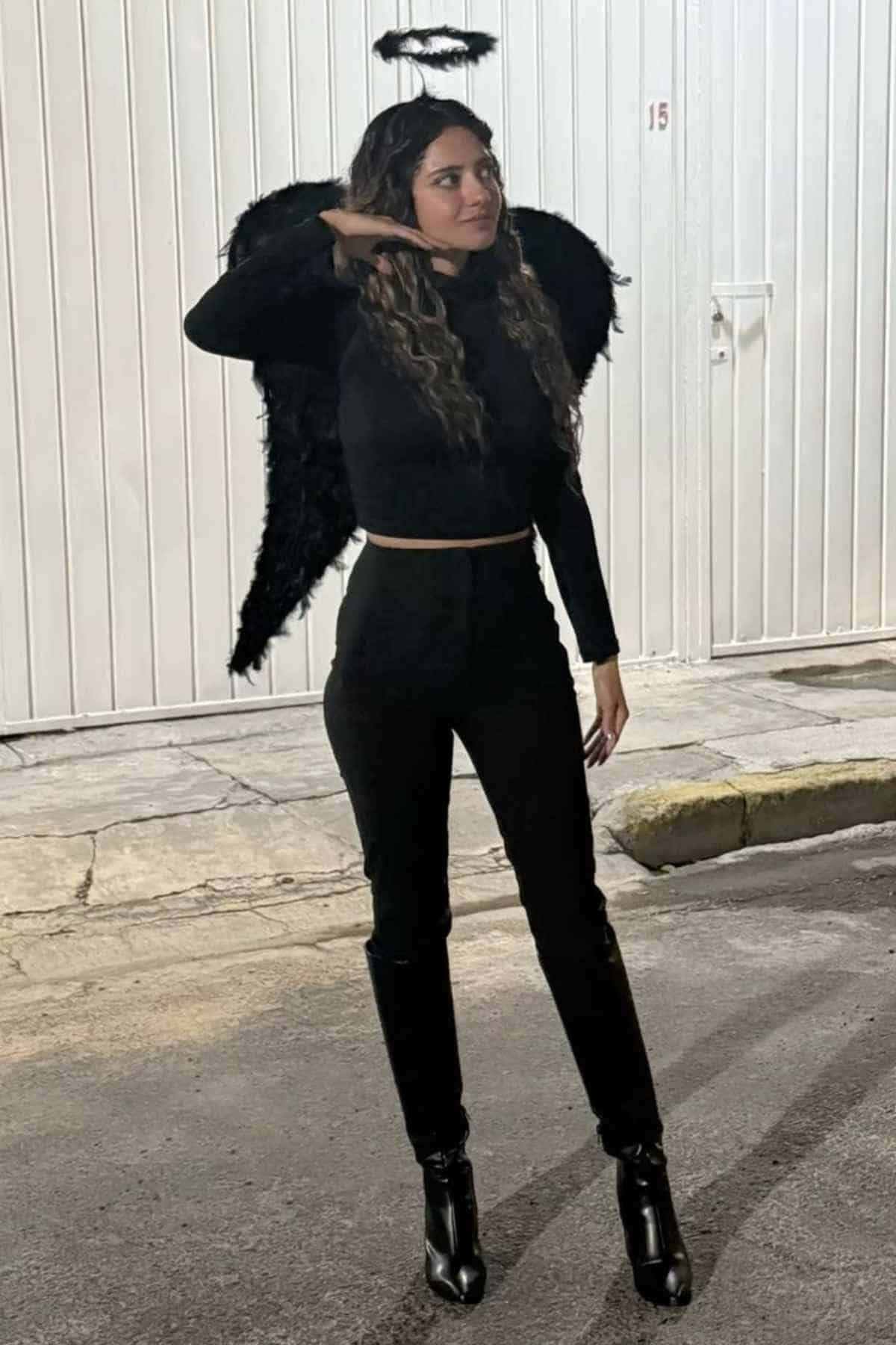 Person wearing a black outfit with black angel wings and a black halo headband, standing on a street at night near a white garage door.
