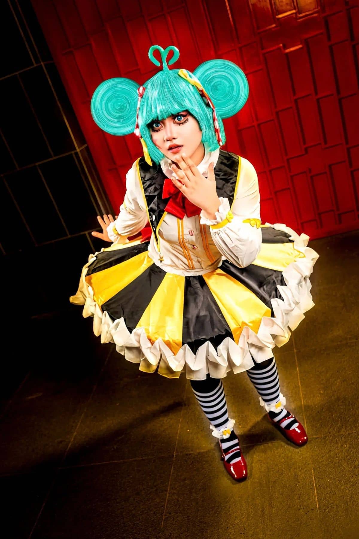 Person in a colorful, whimsical costume with a blue wig, yellow and black dress, striped stockings, and red shoes, standing indoors against a red wall.