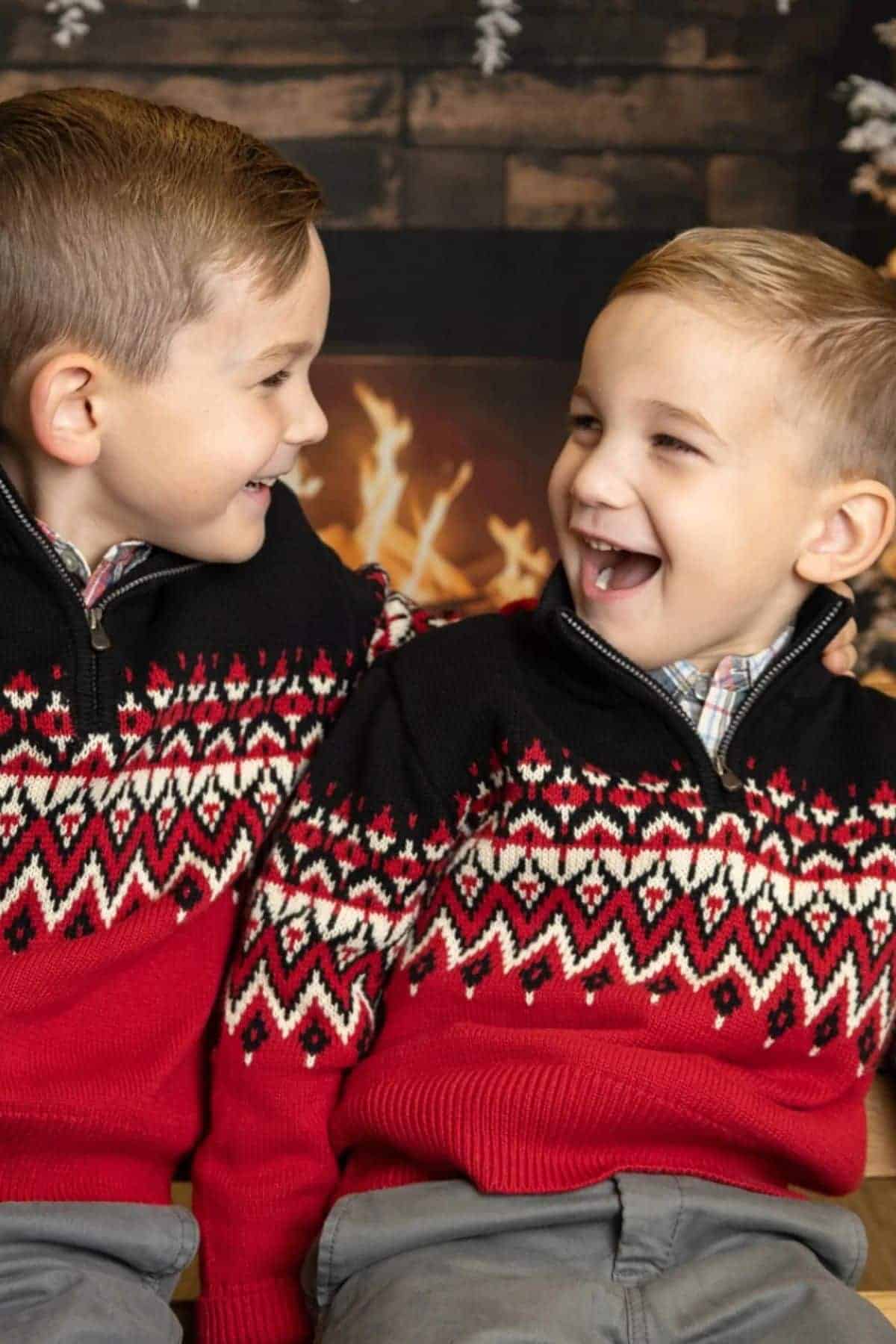 Two young boys in matching red and black patterned sweaters smile and look at each other while sitting in front of a fireplace.