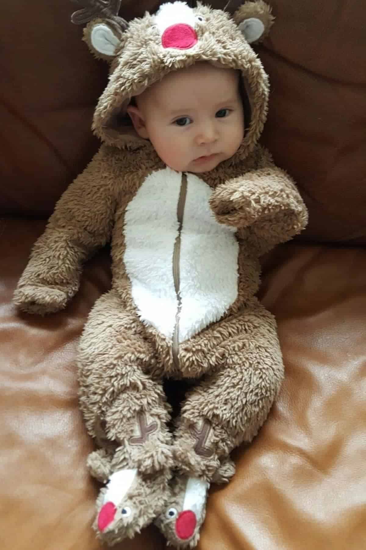 A baby wearing a fuzzy reindeer costume with antlers and a red nose sits on a brown leather couch.