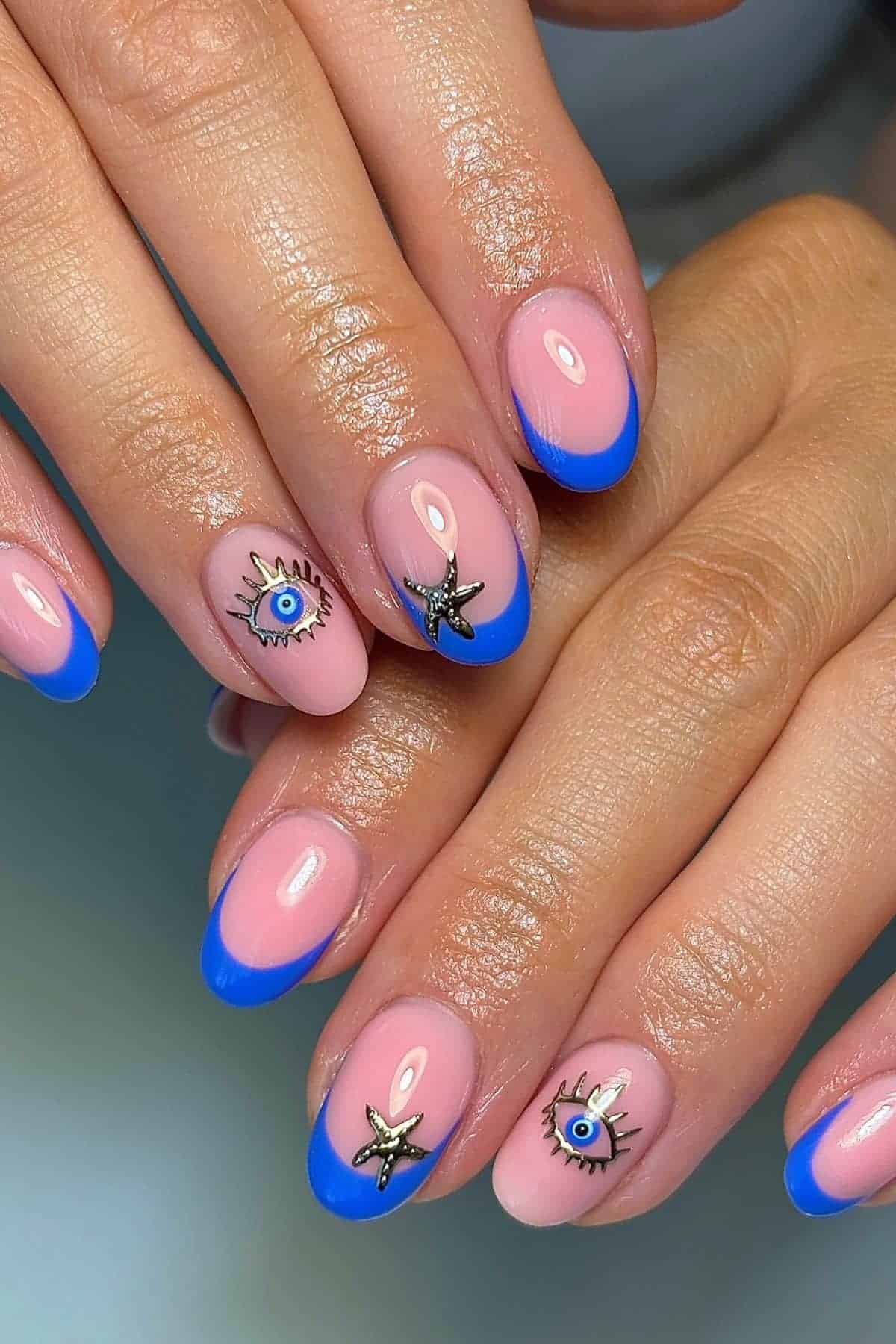 Hands with pink and blue French tip manicured nails featuring gold starfish and blue eye nail art charms on two fingers.