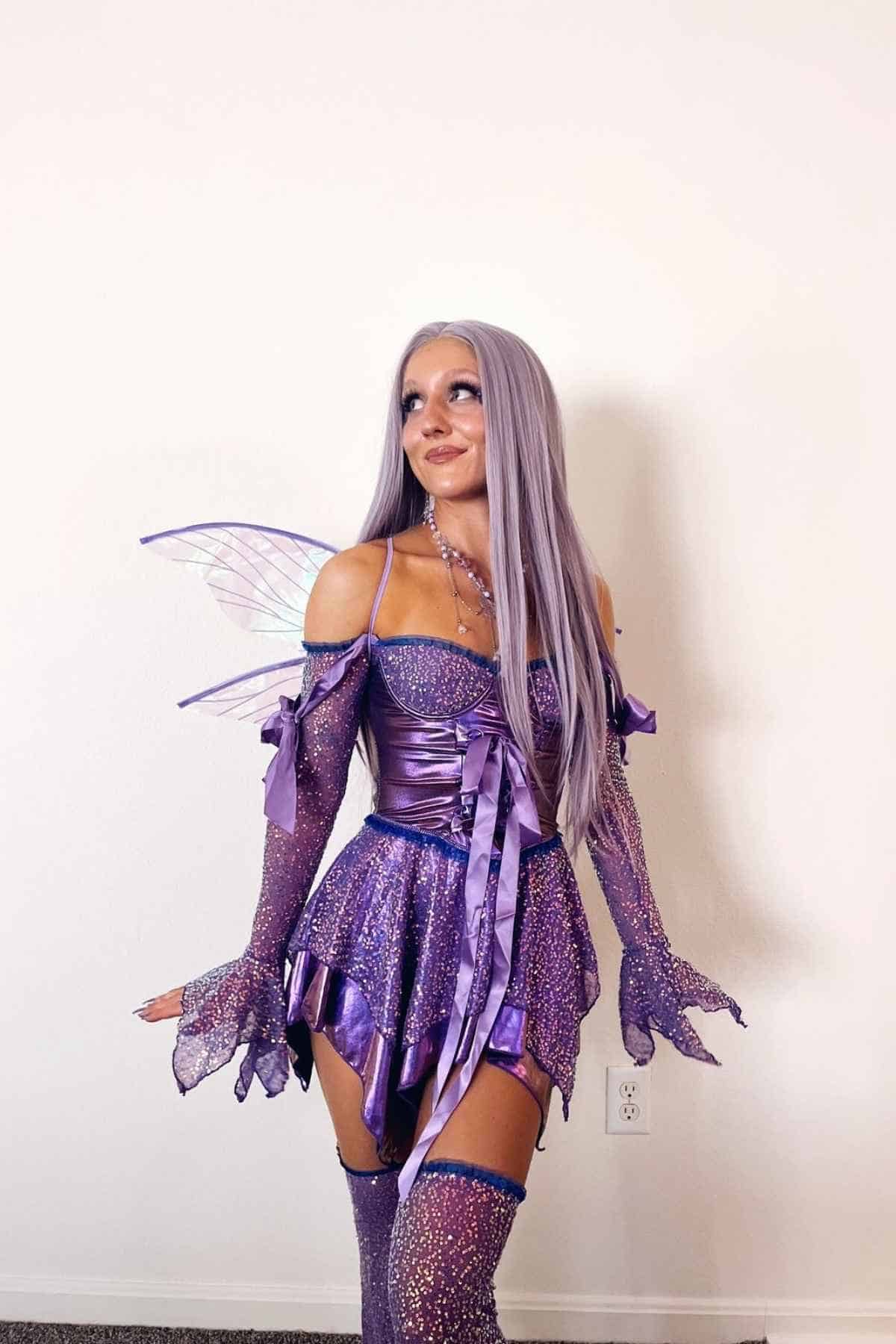Person in a sparkly purple fairy costume with wings, long lavender hair, and gloves, standing against a plain white wall.