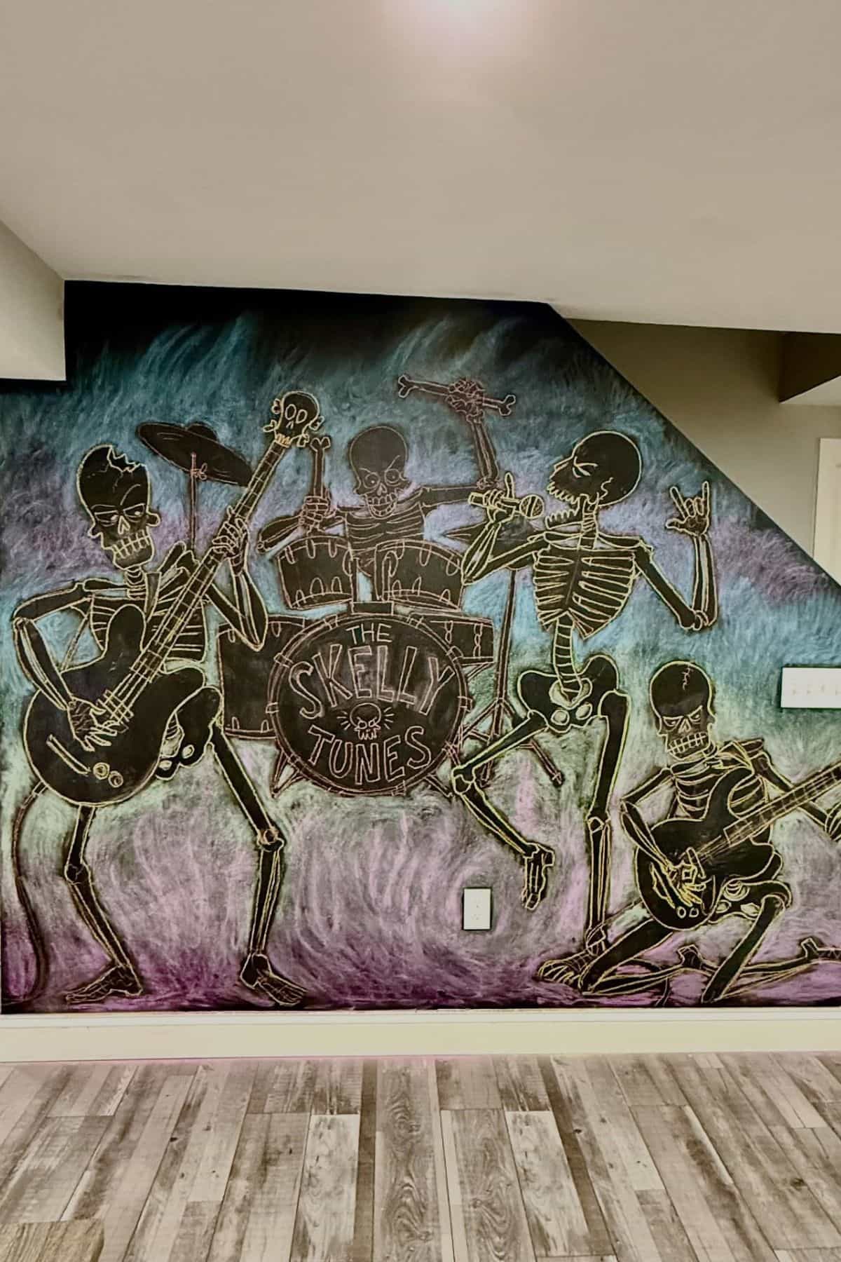 Chalk mural of four skeletons playing instruments as a band called “The Skelly Tunes” on a wall with colorful background.