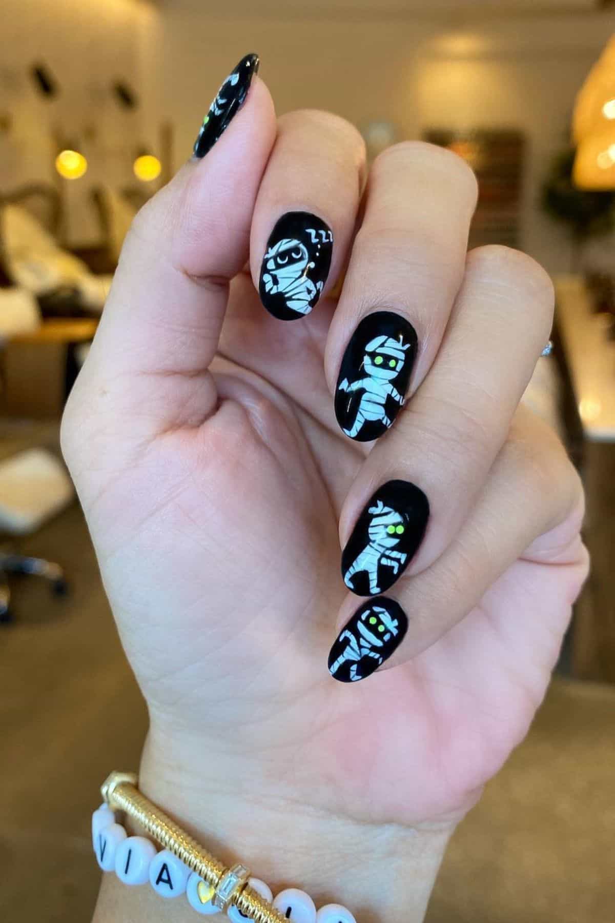 A hand with black nail polish features cartoon mummy designs on each nail. The background appears to be a nail salon with a blurred interior.