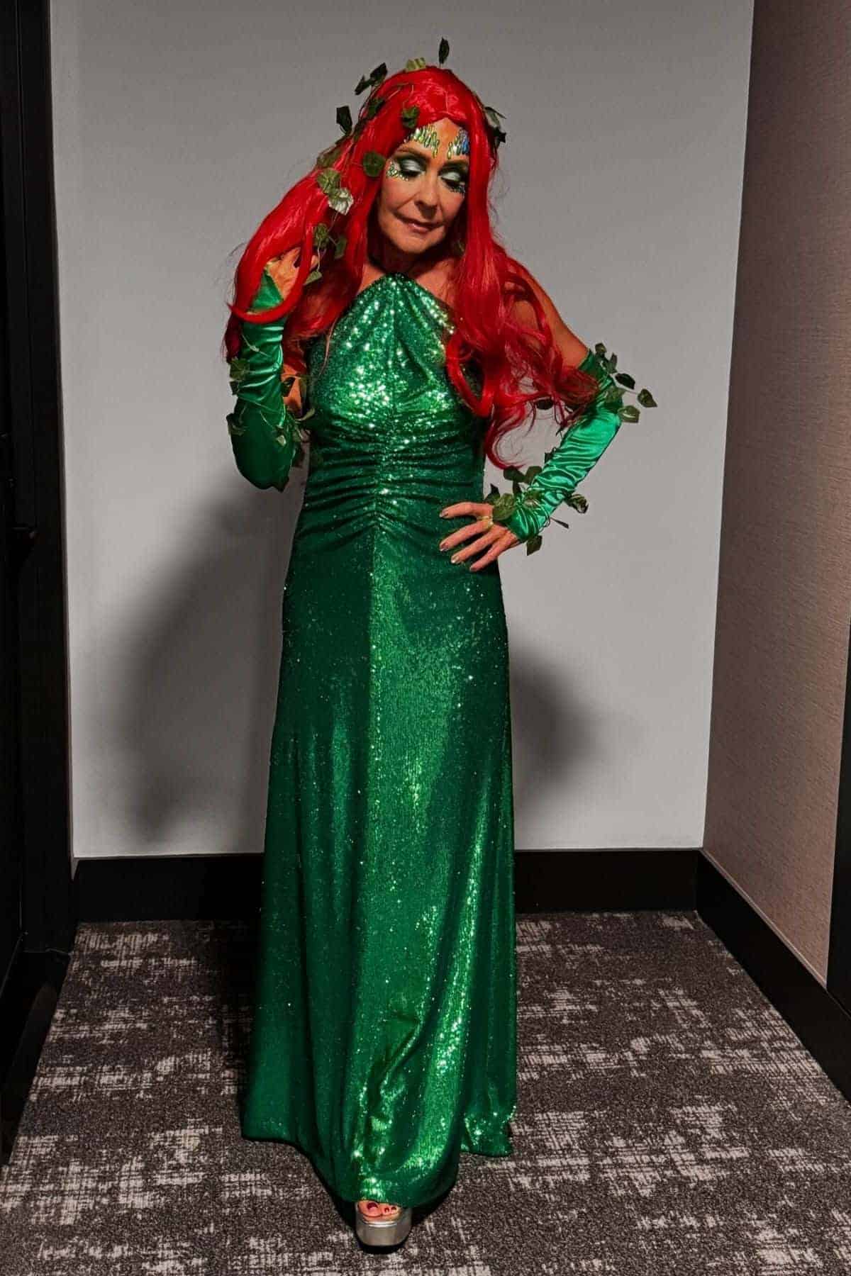 Person wearing a long green glittery dress, red wig, and leaf accessories, standing on a patterned carpet in a hallway.