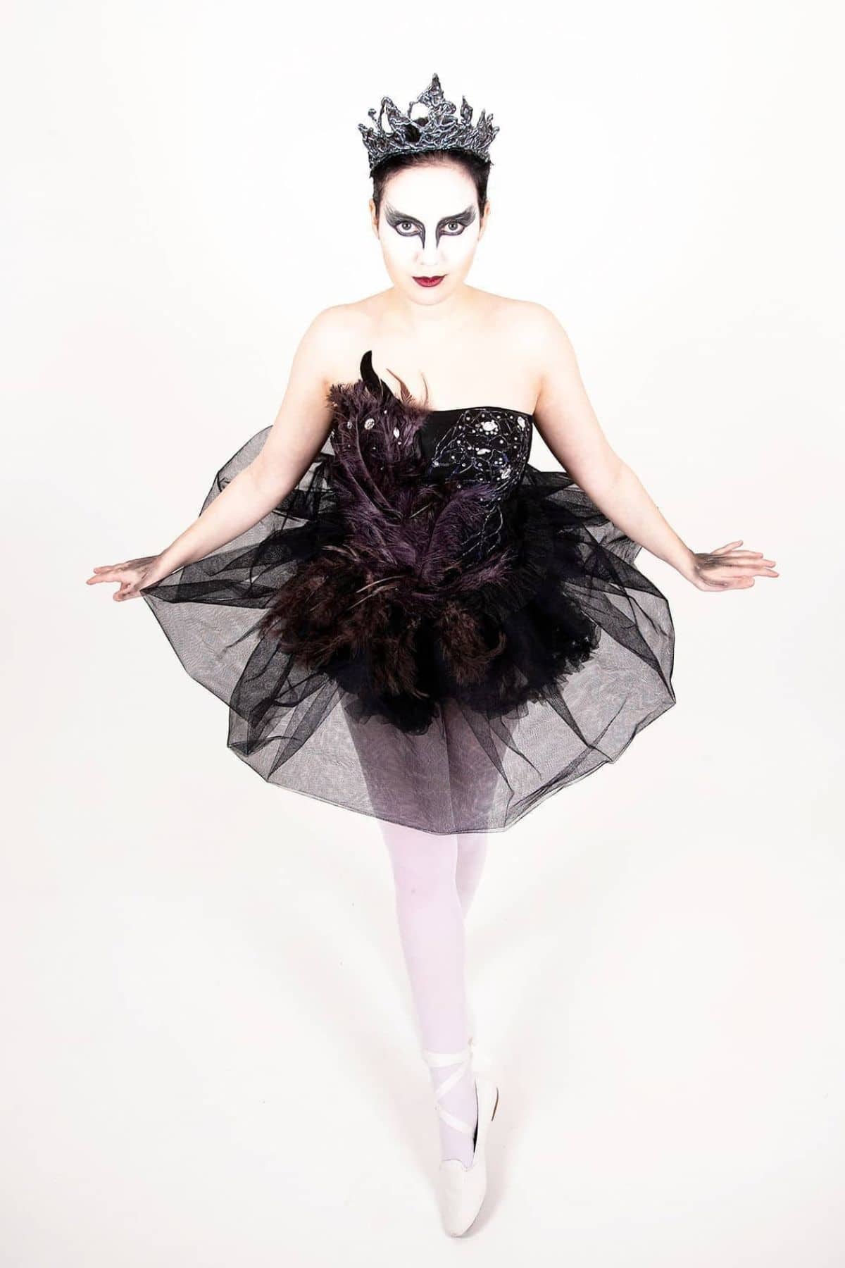 Person in a black, feathered tutu and crown poses en pointe against a white background, with dramatic black-and-white makeup resembling a swan.