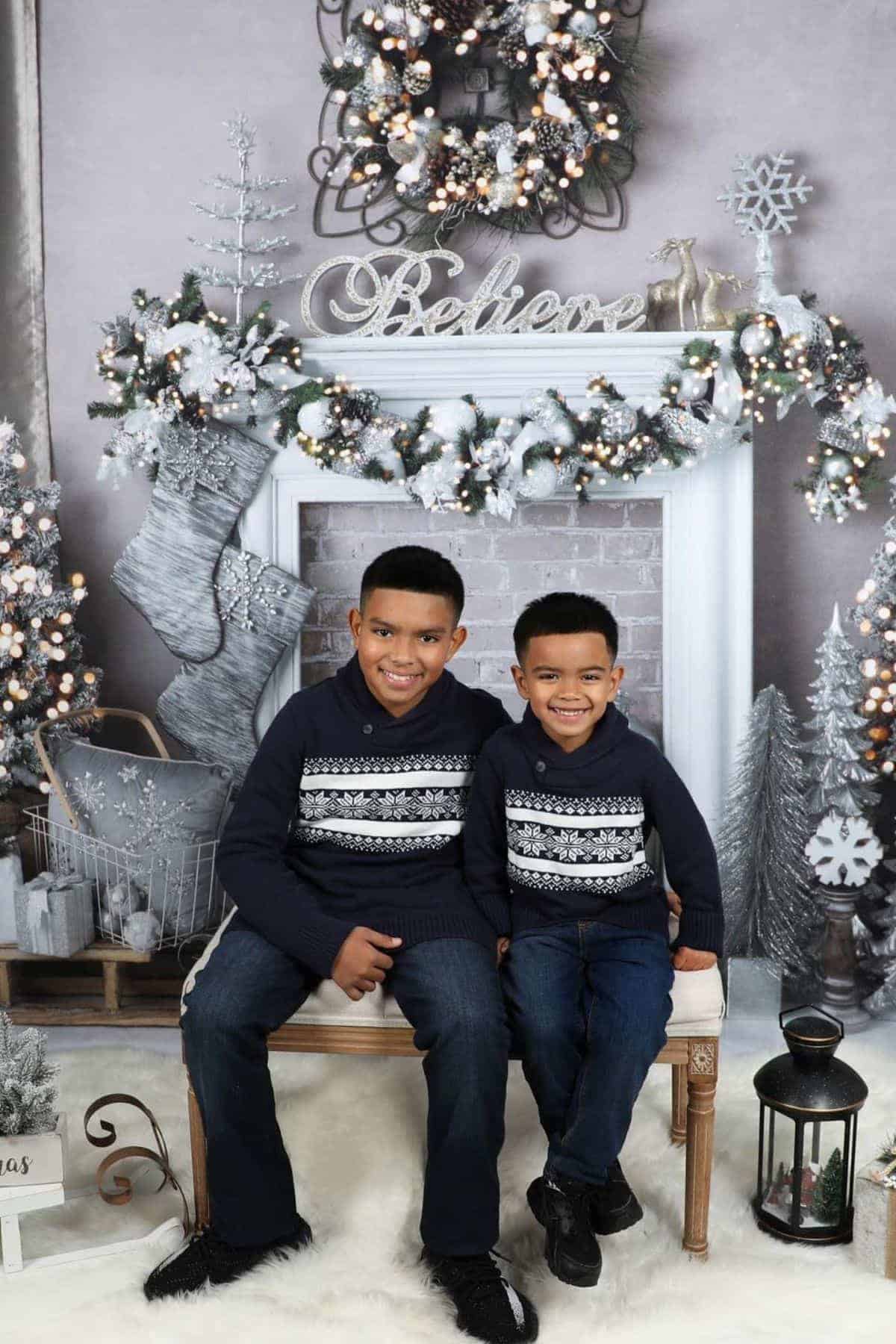 Two young boys in matching sweaters sit on a bench in front of a festive, winter-themed fireplace decorated with garlands, stockings, and ornaments.