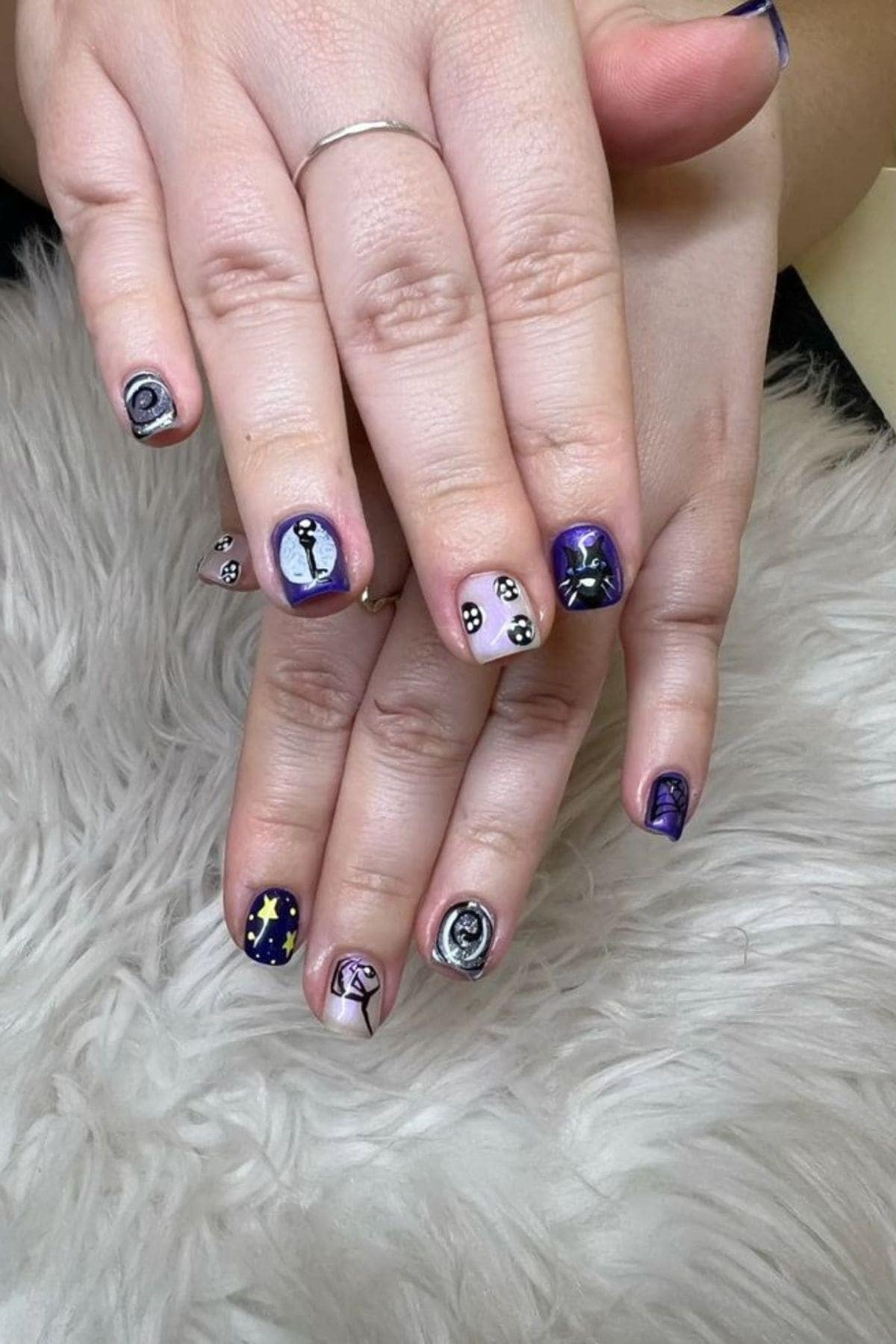 Hands with short nails featuring galaxy-themed and cat nail art designs, including stars, moons, and cat faces, resting on a shaggy white surface.