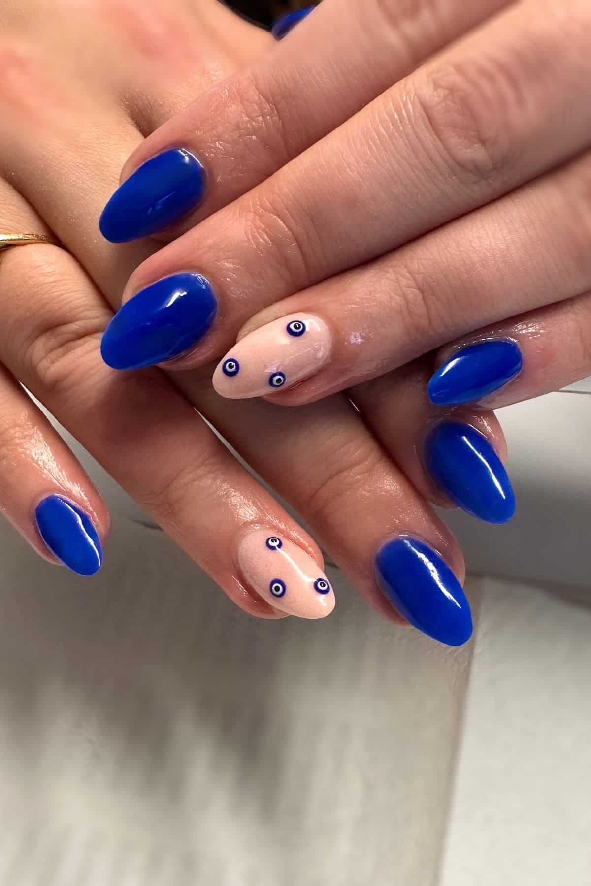 Hands with blue oval-shaped nails; ring fingers have nude polish with small blue and white eye-like designs.