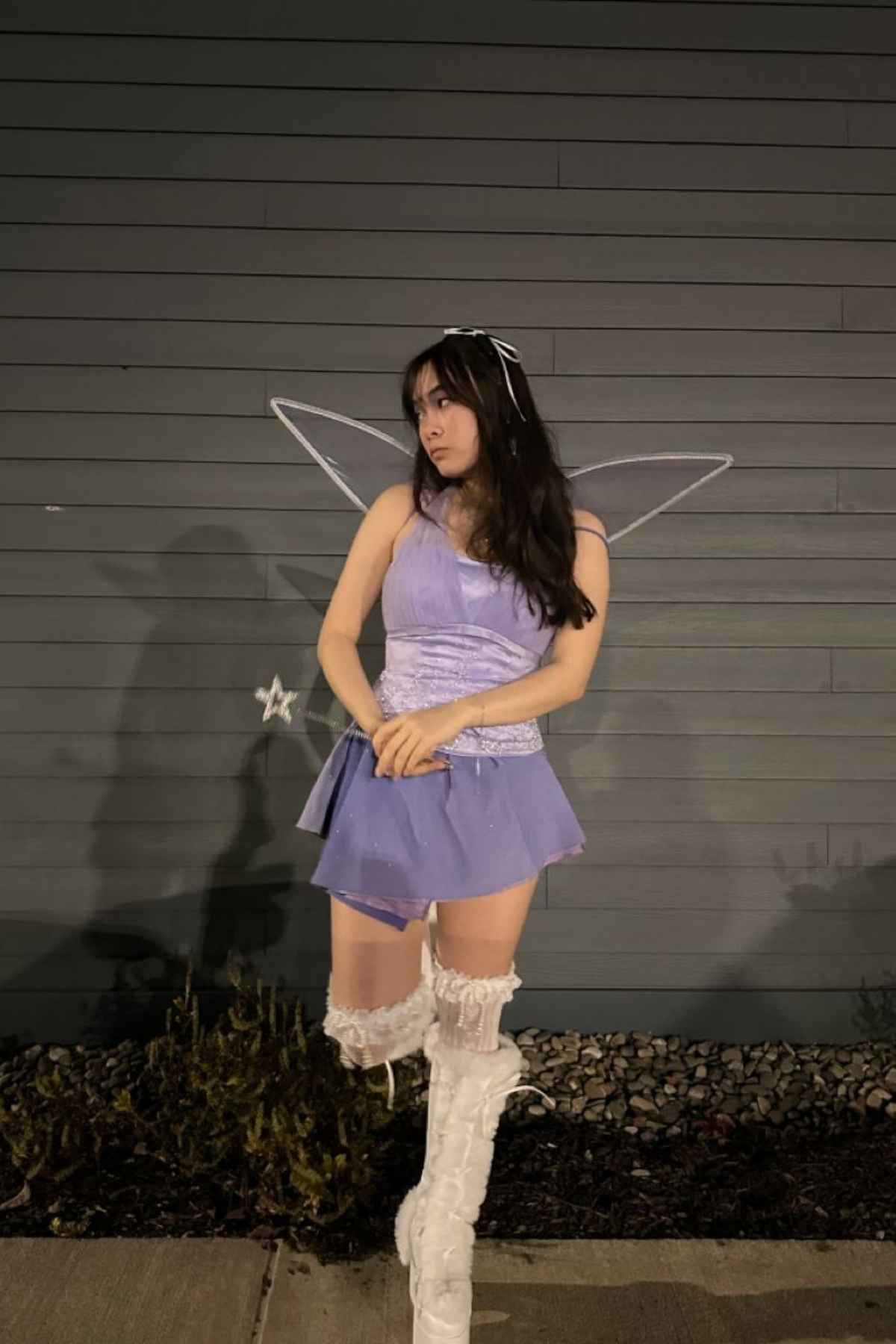 A person in a lavender fairy costume with wings and white boots stands in front of a gray wall at night.