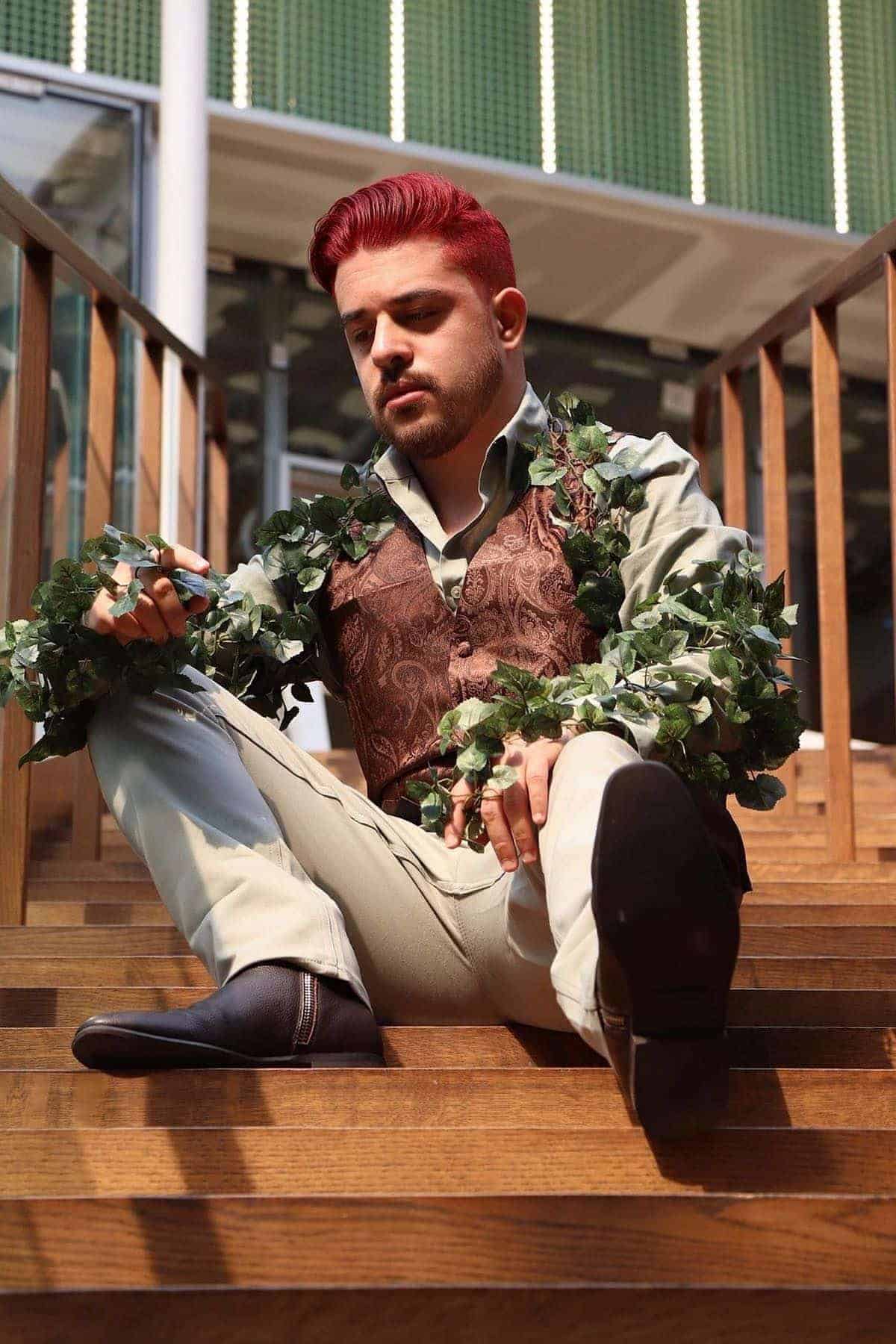 A person with red hair sits on wooden stairs, wearing light pants, a patterned brown vest, and a shirt adorned with leafy vines, holding a phone and looking down.