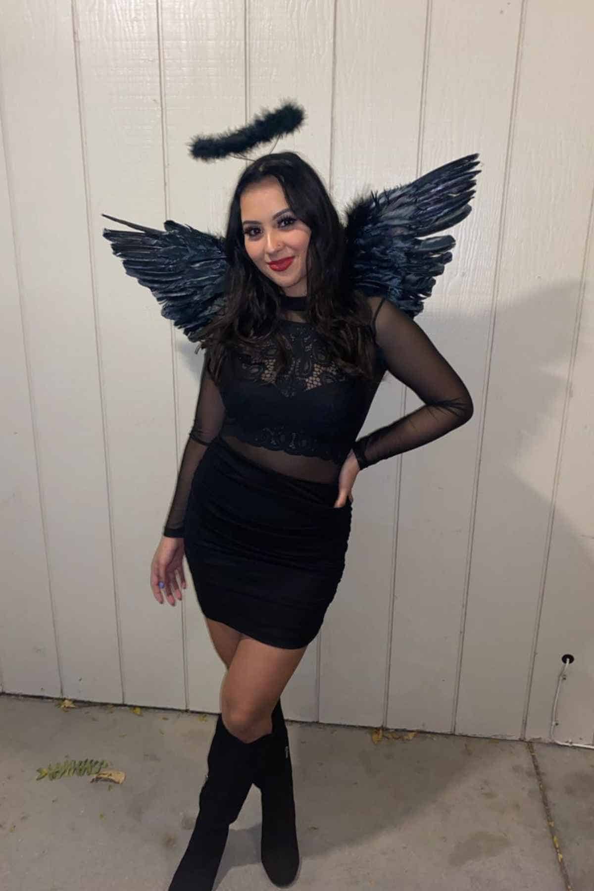 Woman poses in front of a beige wall wearing a black halo headband, black feathered wings, a black sheer top, black skirt, and black boots.
