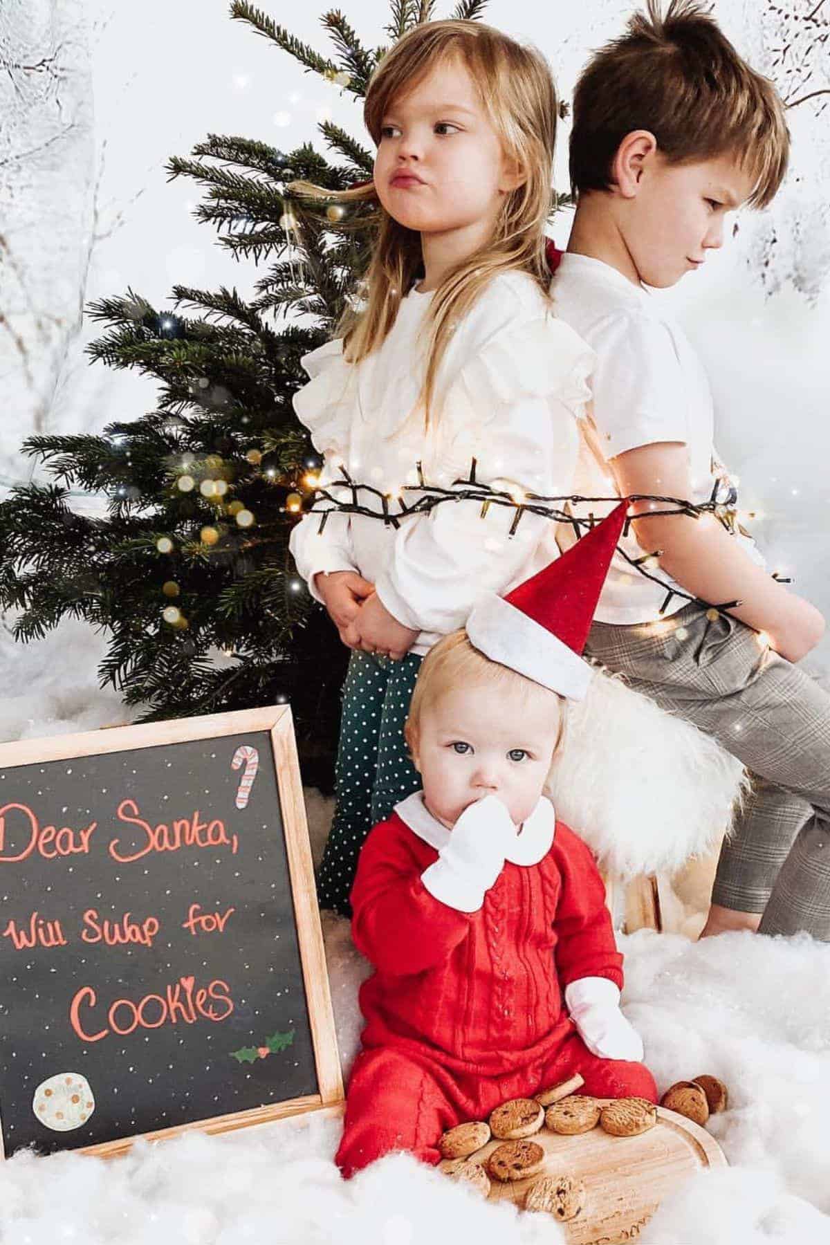 Three children sit in front of a Christmas tree; one is dressed as Santa. A chalkboard beside them reads, “Dear Santa, Will swap for cookies.” String lights and cookies are also visible.
