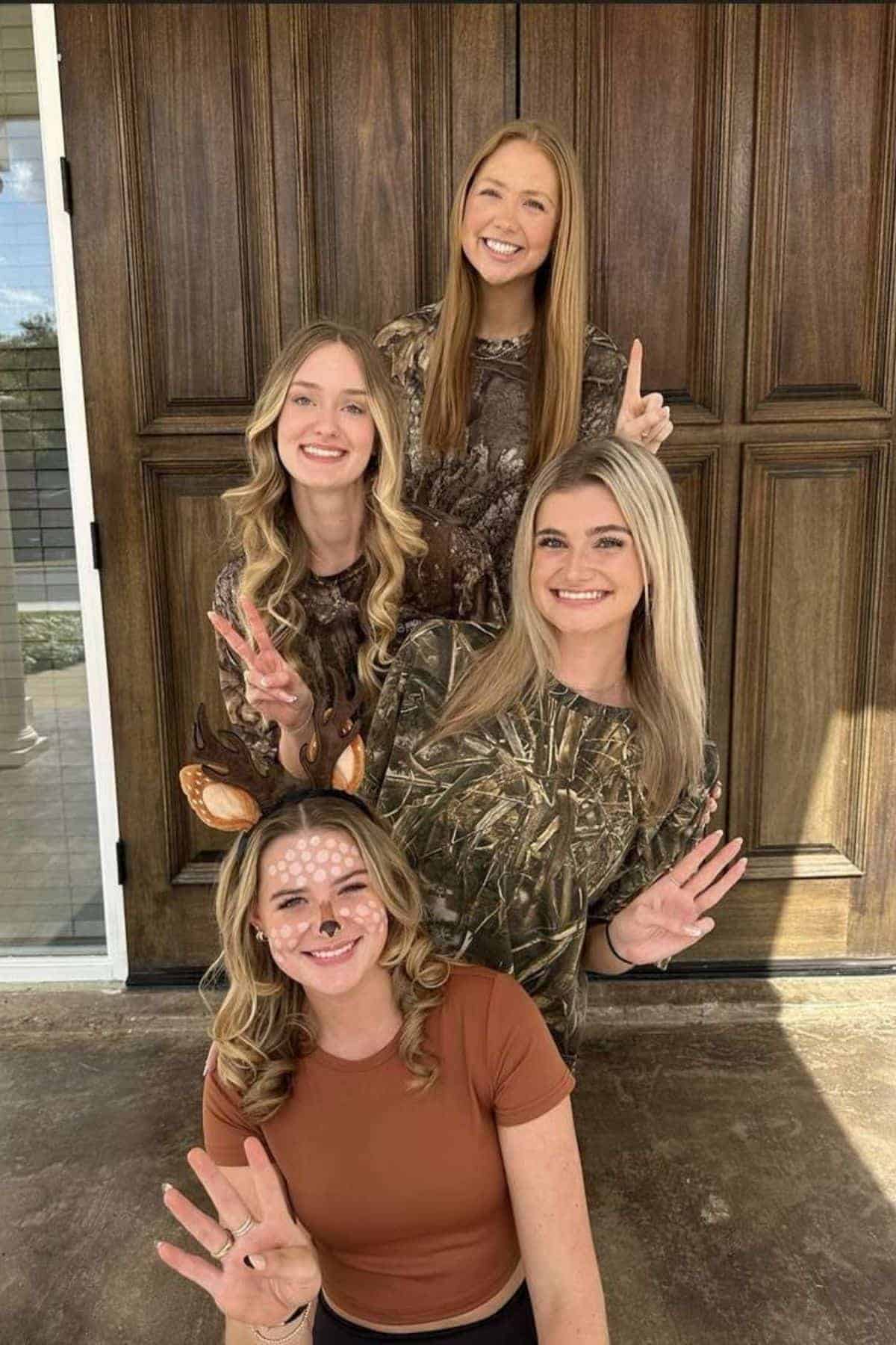 Four women pose in front of a wooden door; three wear camouflage shirts and hold up fingers, while one in front wears antlers and has her face painted like a deer.