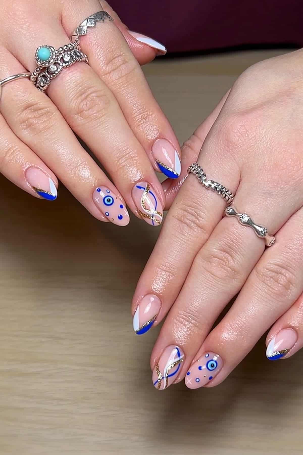 Hands with silver rings display manicured nails featuring blue tips, gold accents, and evil eye designs on a wooden surface.
